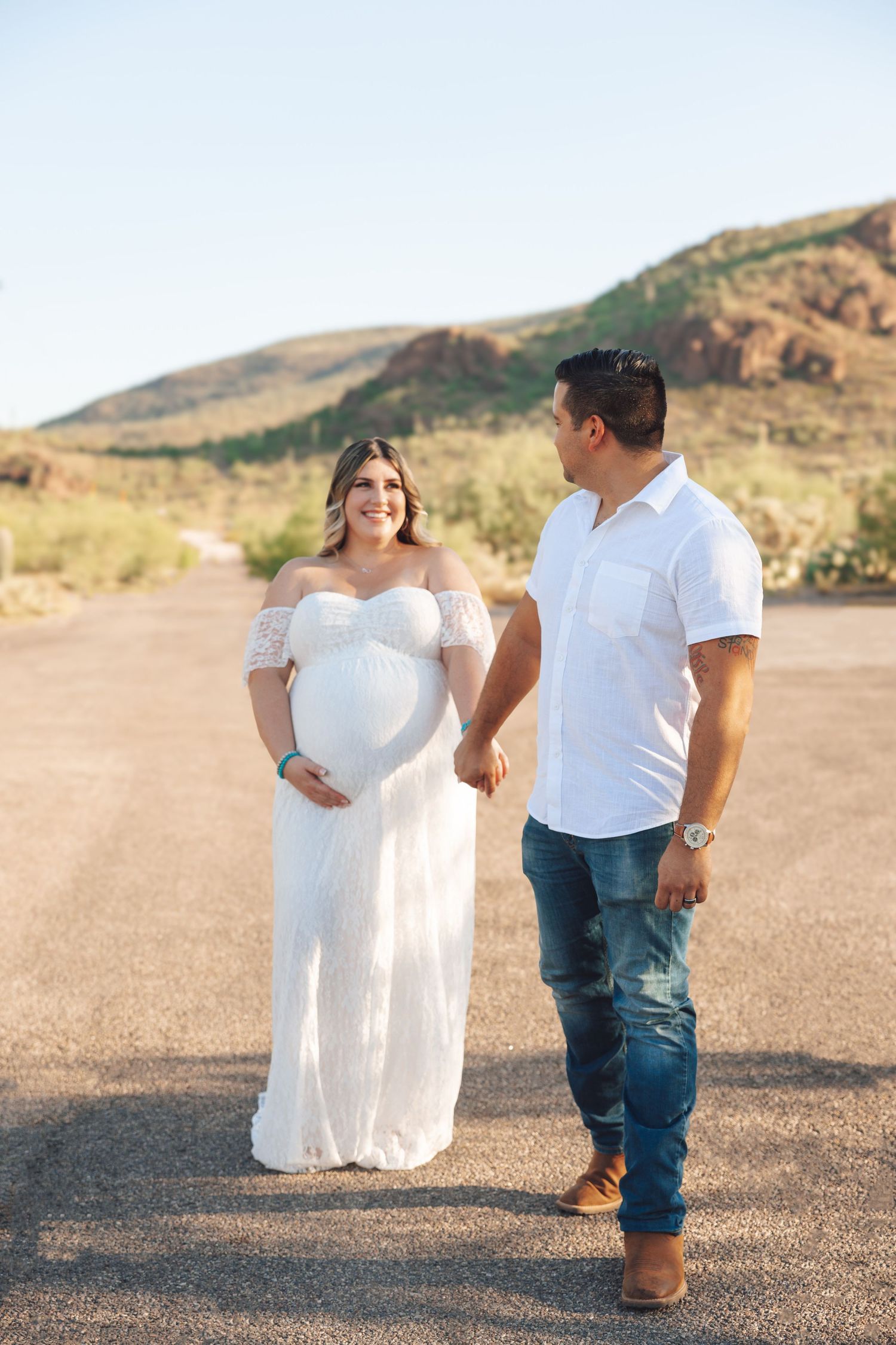 A couple stands together on a desert road with mountains in the background during a maternity photoshoot.