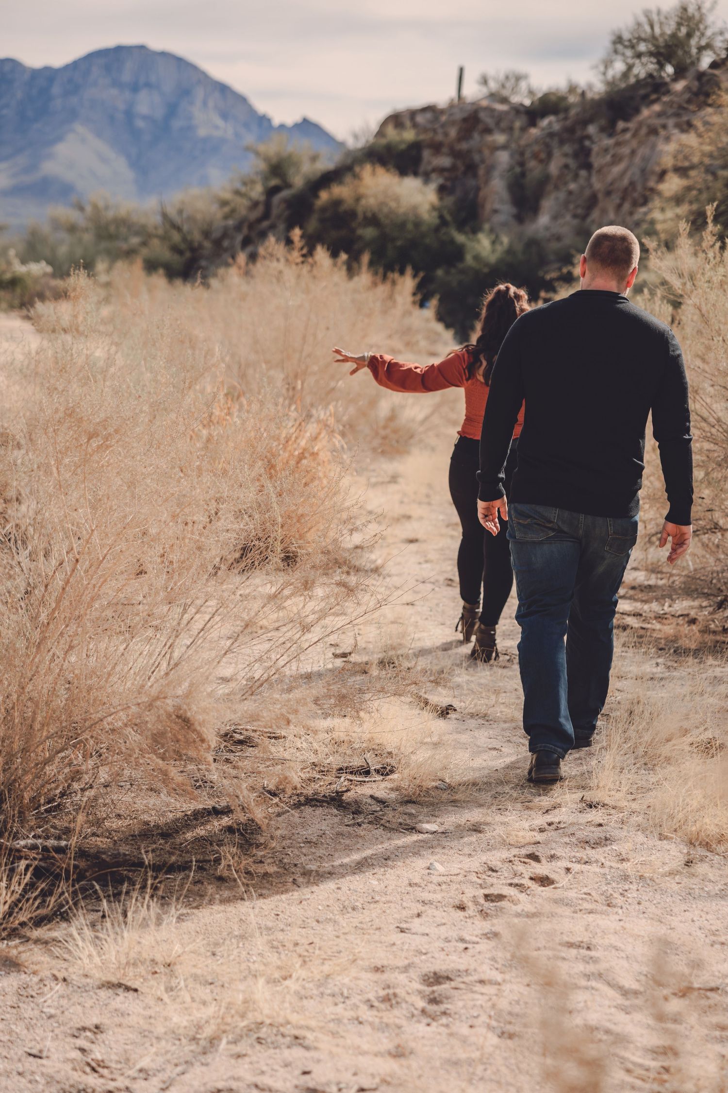 People walking on a dusty desert trail with dry brush and mountains in the background.