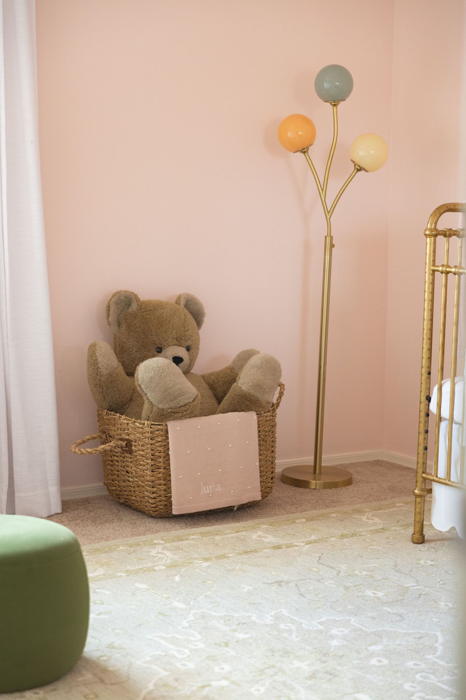 A cozy nursery corner with a teddy bear in a wicker basket, gold floor lamp, and pink walls.