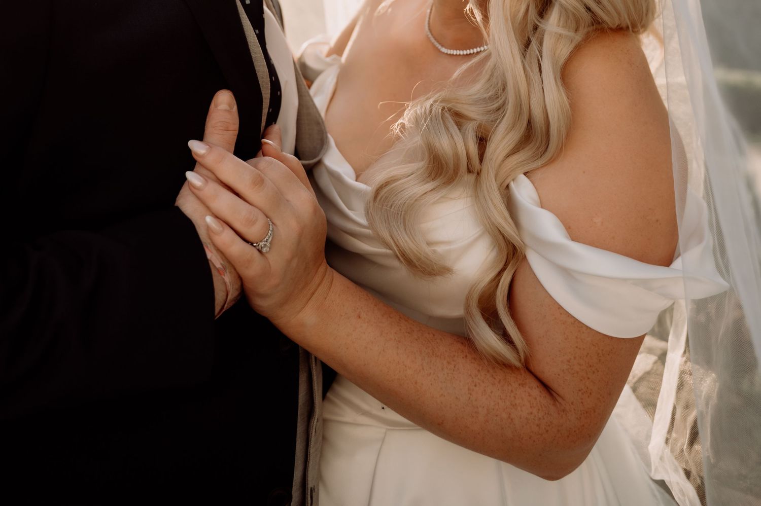 Bride in white off-shoulder gown embraces groom, showcasing diamond engagement ring and flowing veil.