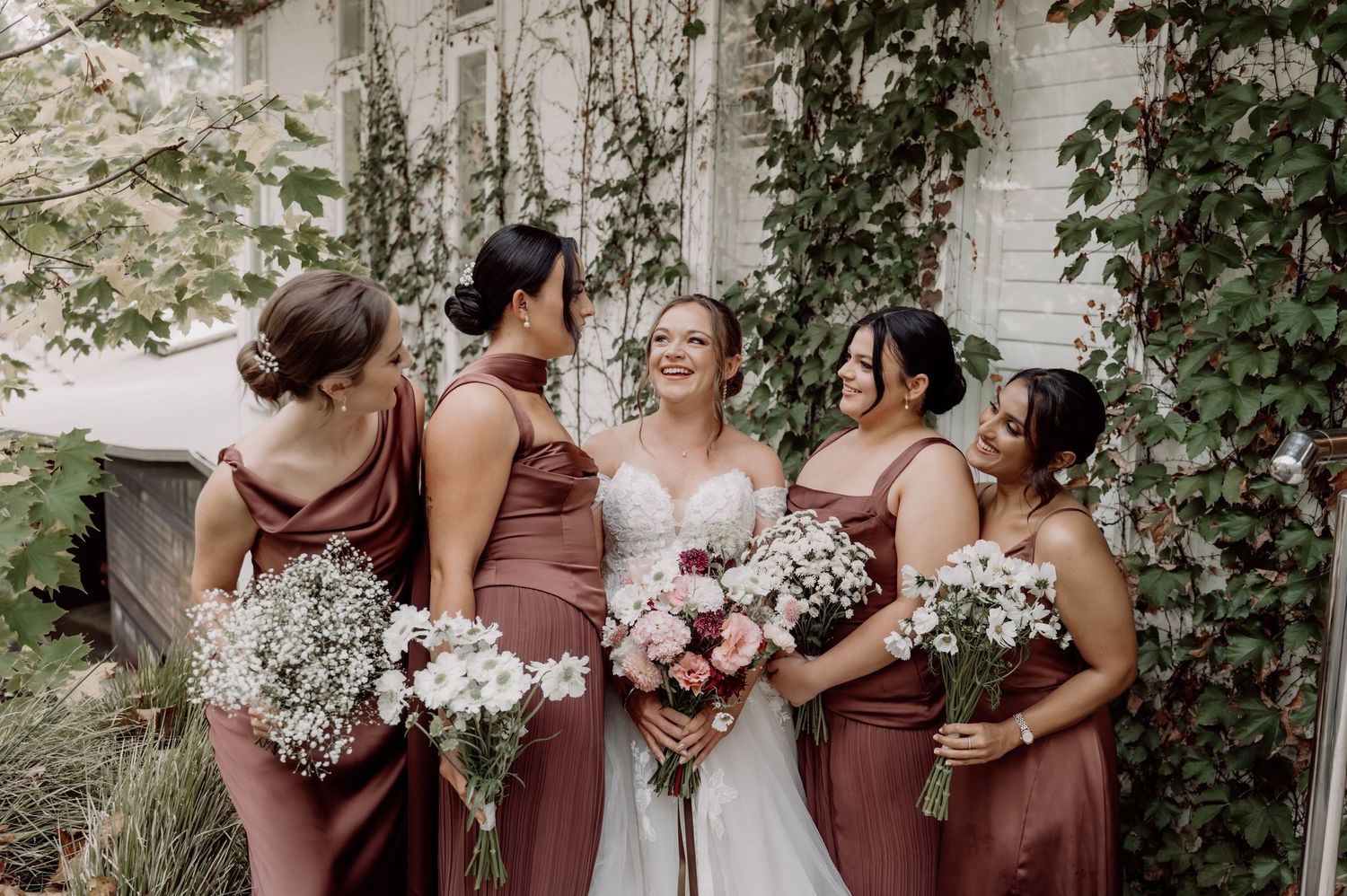 Bride in white gown with three bridesmaids in dusty rose dresses holding bouquets, laughing outdoors near ivy-covered building.