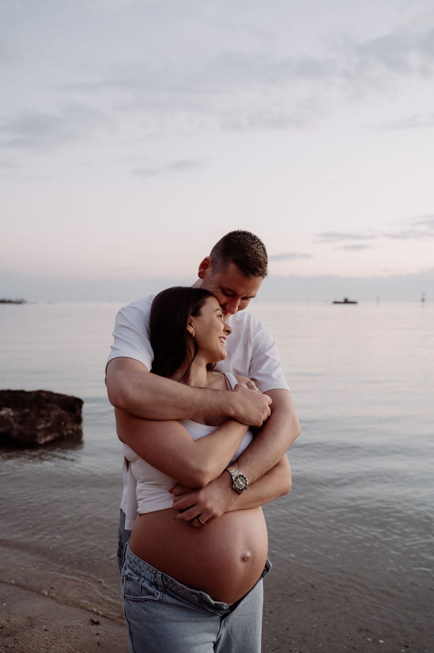 Couple embracing on beach at sunset, showing pregnant belly, man in white shirt hugging smiling woman.