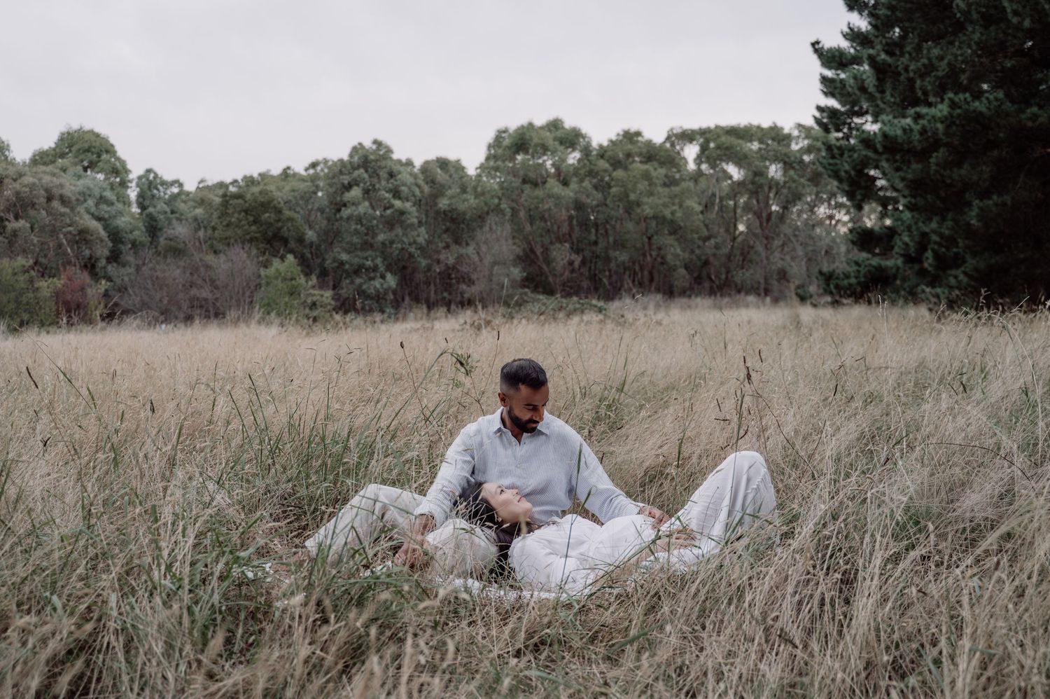 Couple sitting together in a golden grass field with lush green trees in the background on an overcast day.