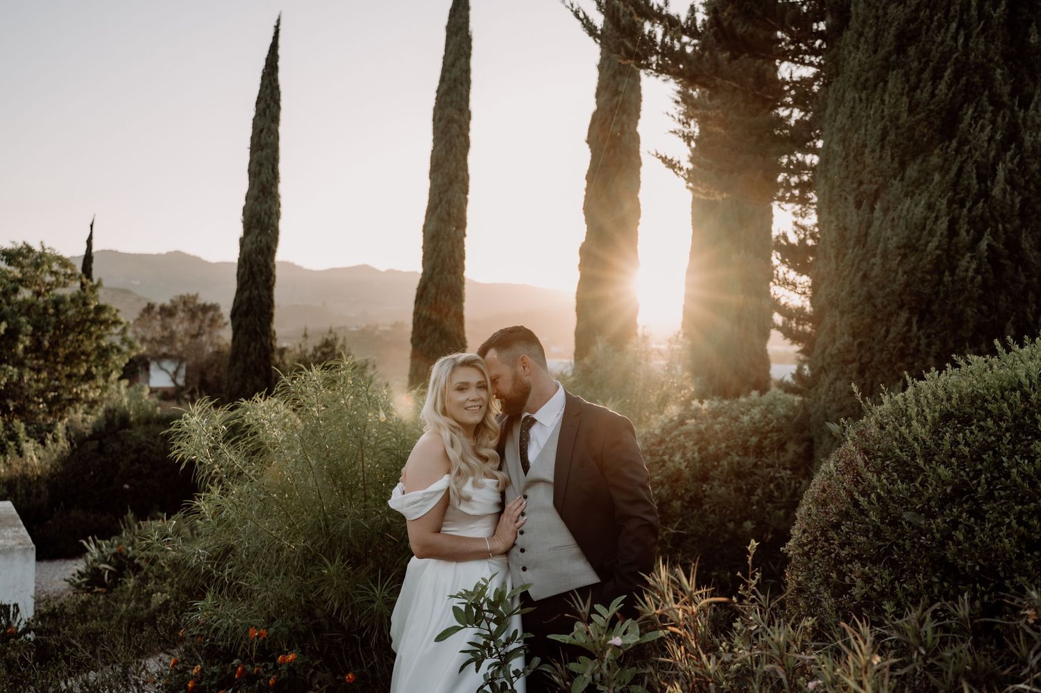 Couple embraces at golden hour sunset in lush garden with tall cypress trees and mountain backdrop.