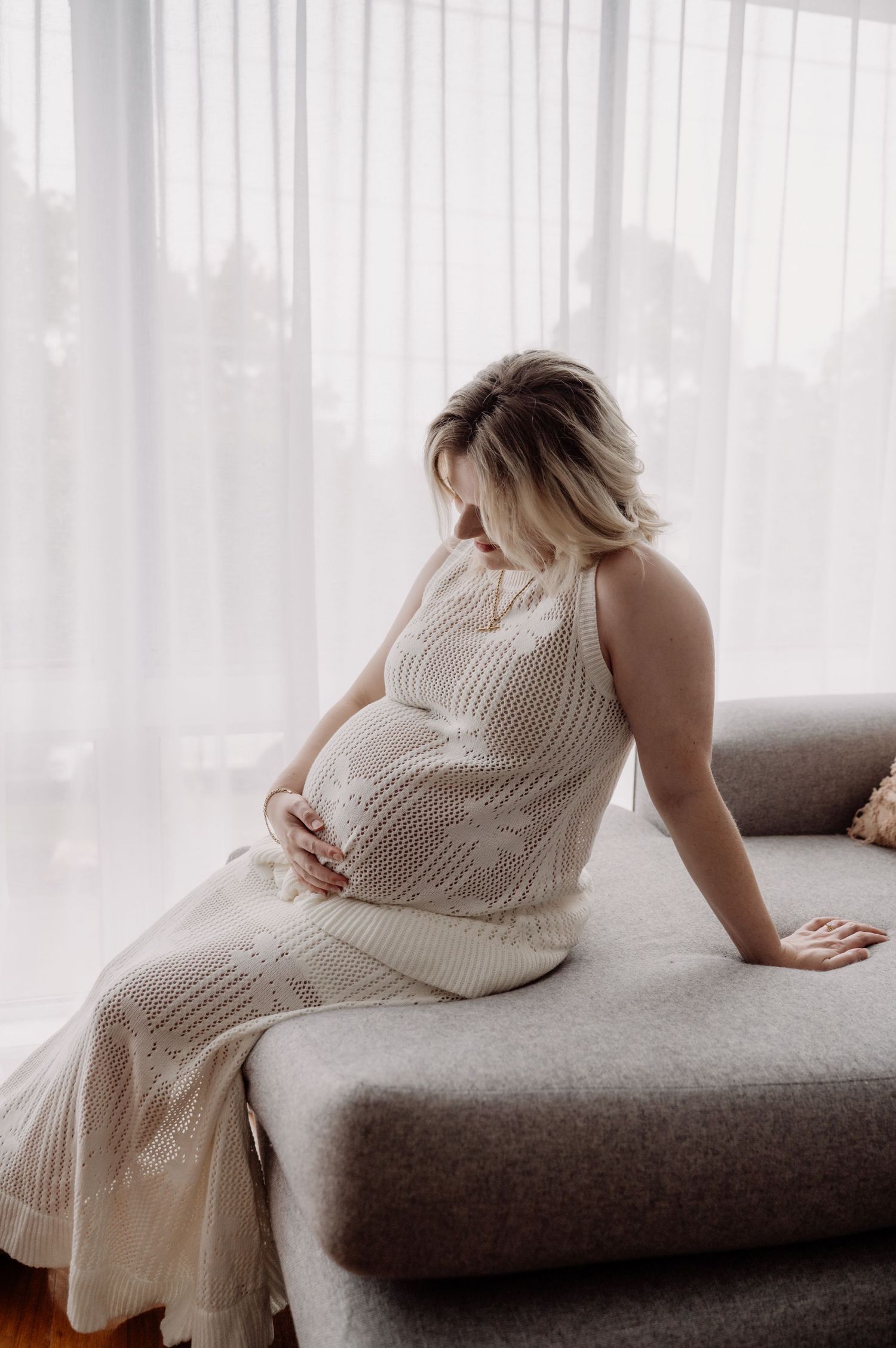 Pregnant woman in white knit dress sitting on gray sofa, cradling her baby bump near sheer curtains.