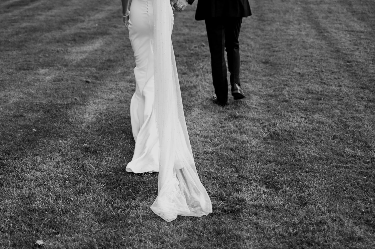 Black and white photo of bride in flowing wedding gown walking with groom on grass.