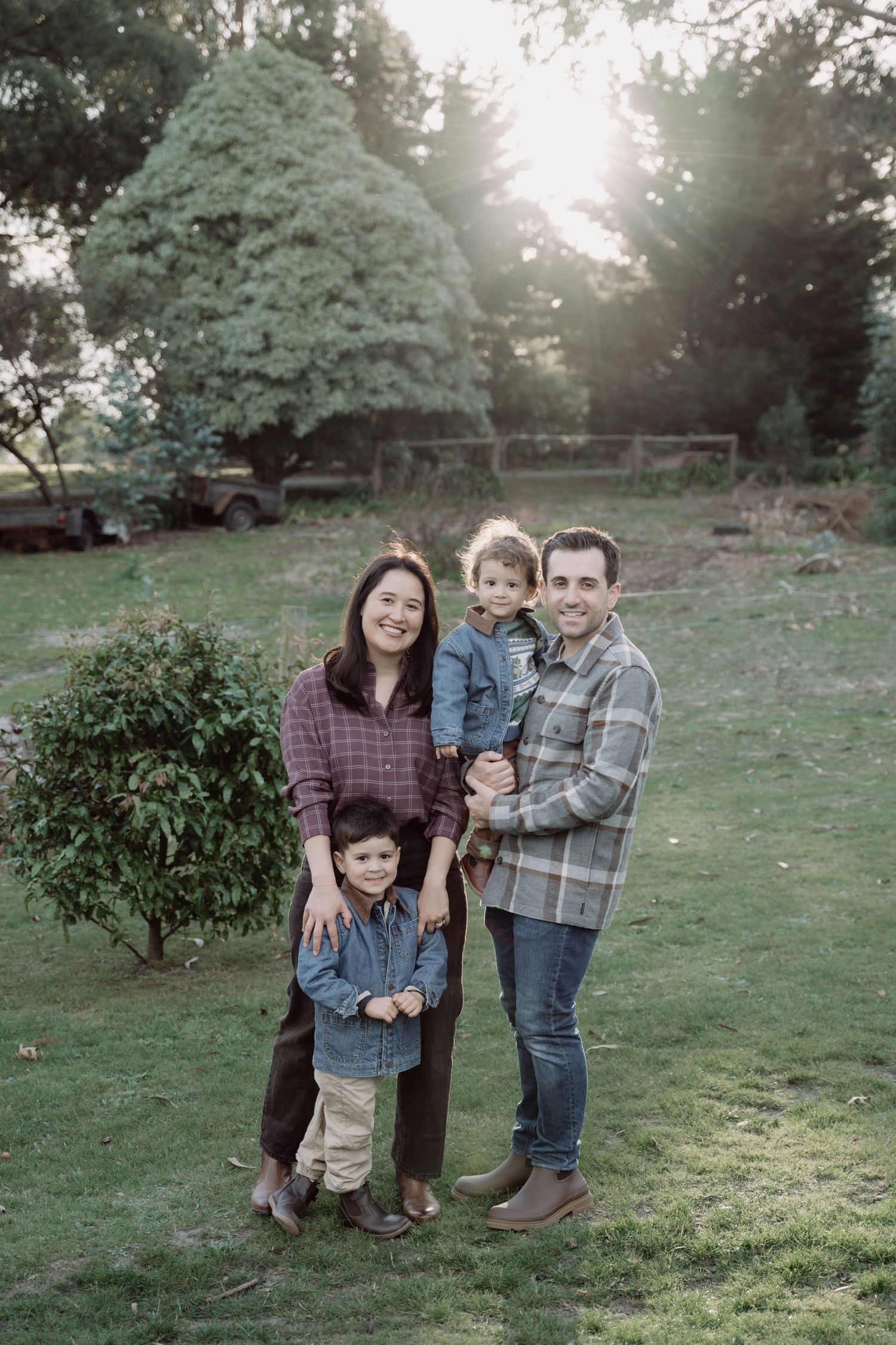 Family of four posing outdoors at golden hour on green lawn, parents and two young children smiling, trees in background.