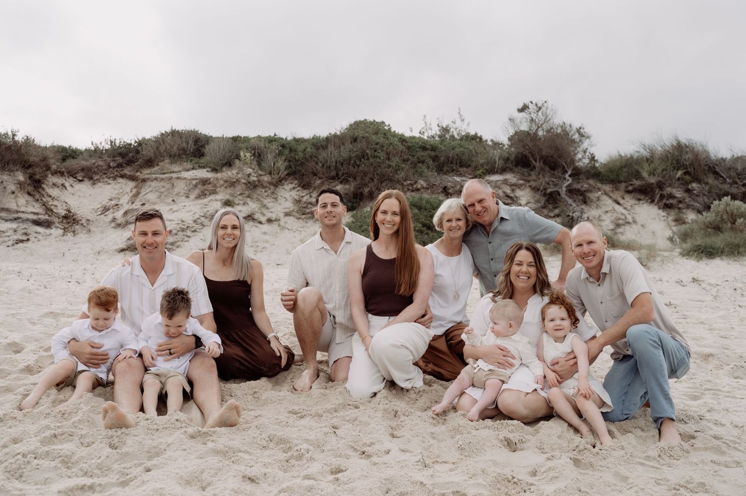 Large family group portrait on sandy beach with cloudy sky, adults and children smiling together near coastal dunes.