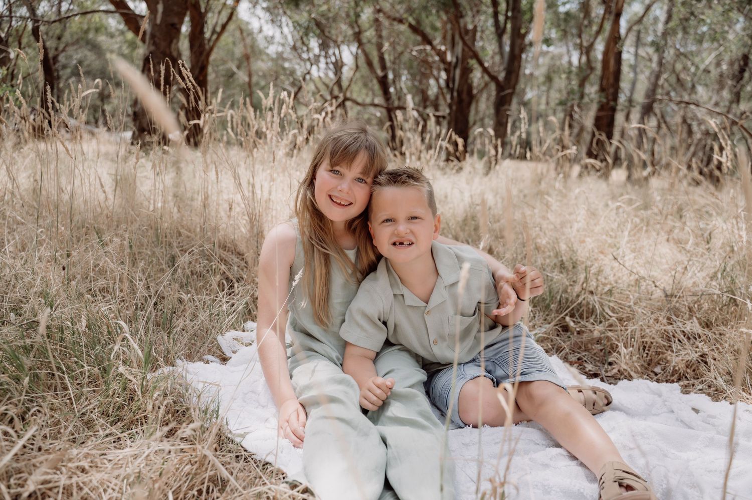 Two smiling children sitting together on a blanket in a natural Australian bush setting with golden grass and eucalyptus trees.