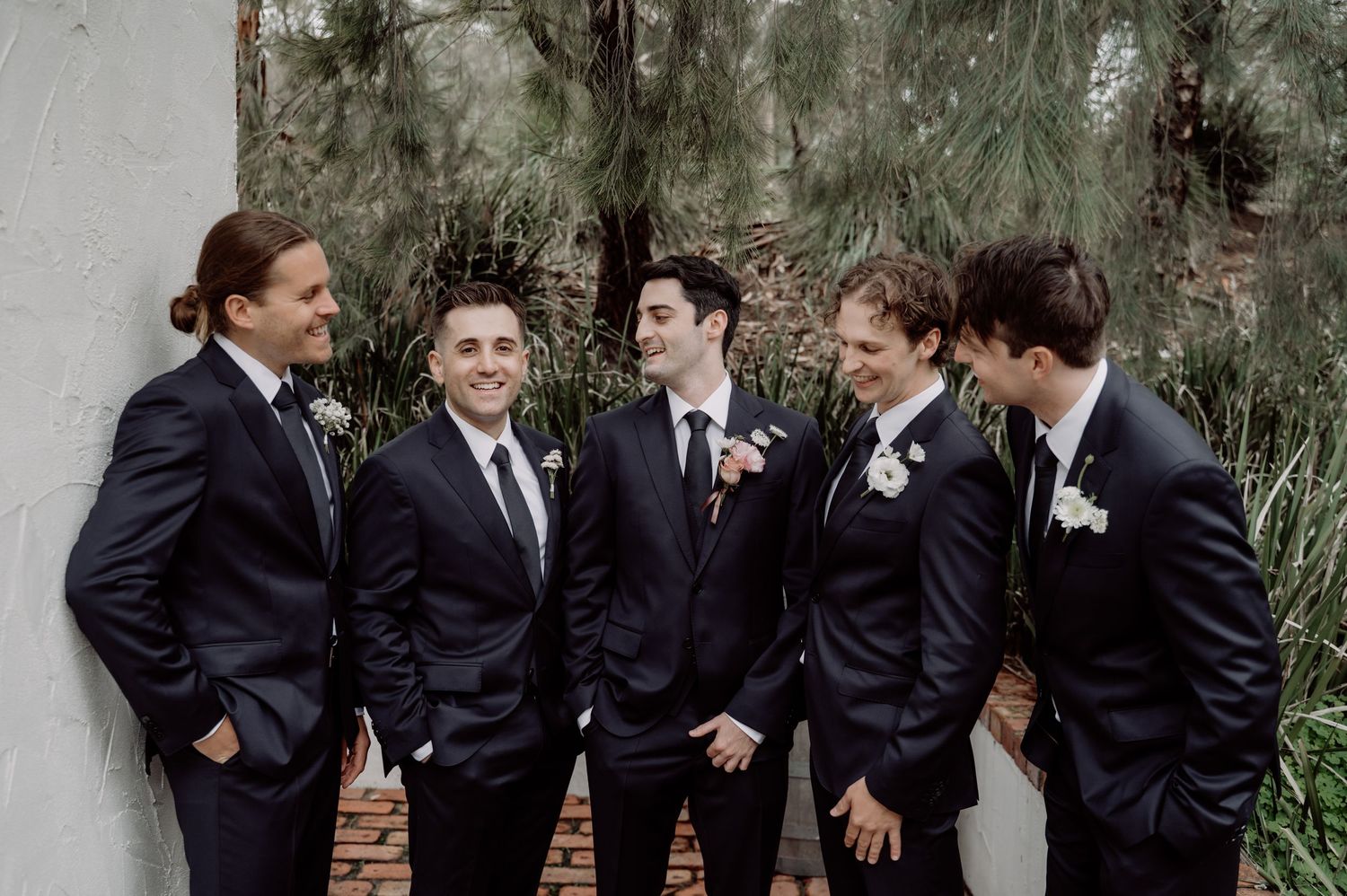 Five groomsmen in navy suits with white boutonnieres laughing together outdoors at a wedding celebration.