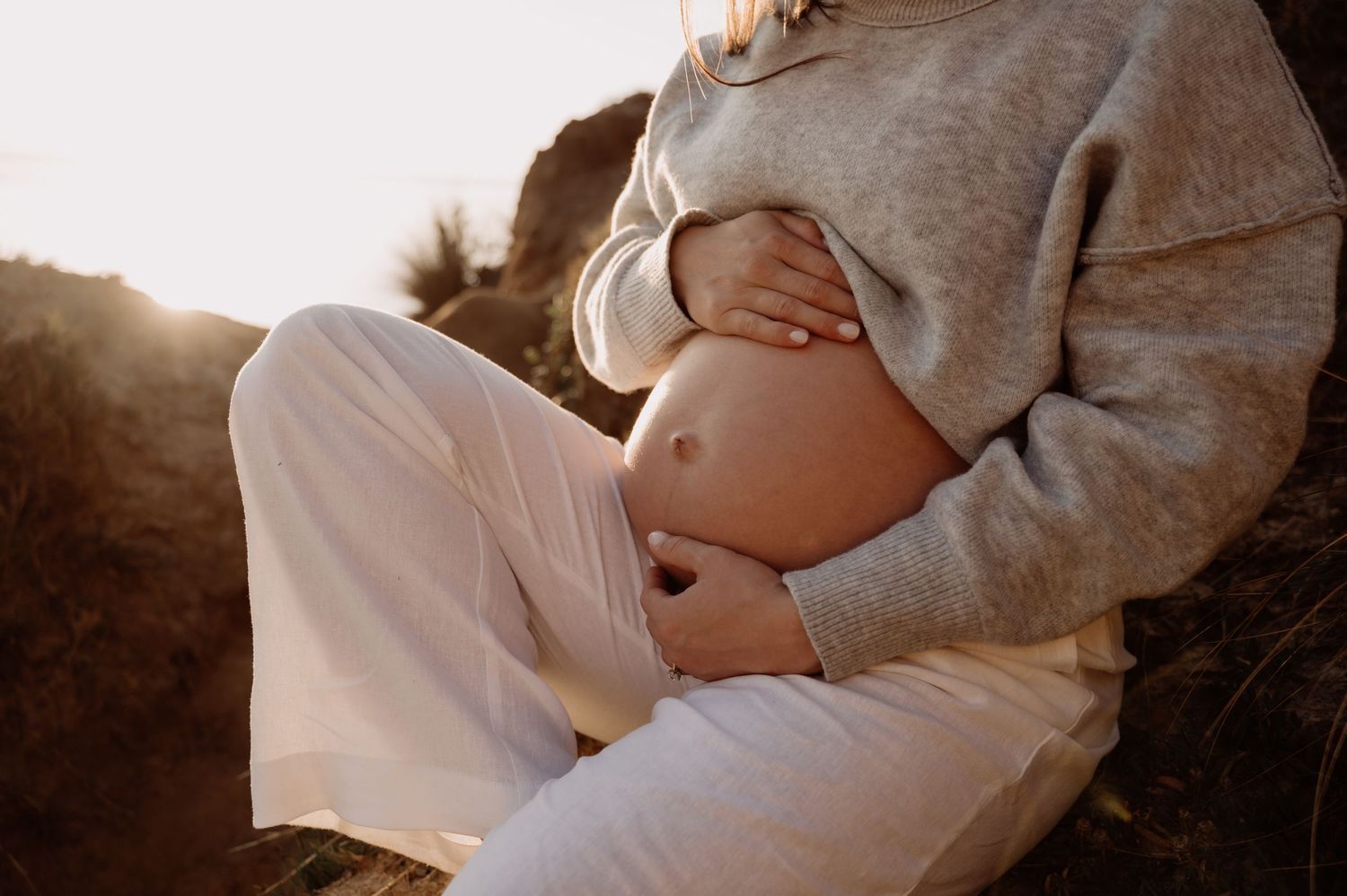 Pregnant woman in gray sweater and white pants cradling her baby bump outdoors in golden sunlight.