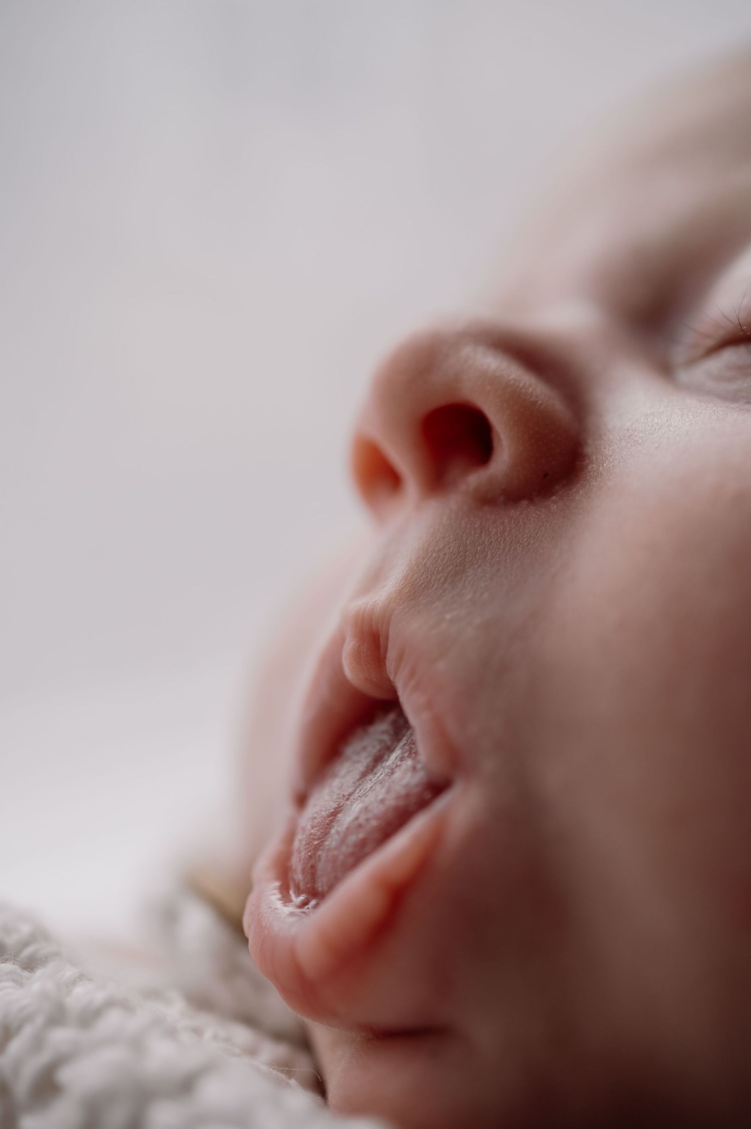 Close-up of a sleeping newborn baby with mouth open and tongue out, resting on a soft white blanket.