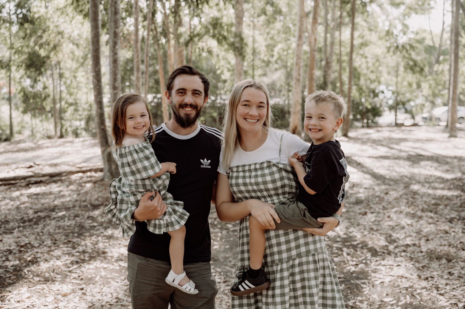 Happy family of four smiling outdoors in a forest, parents holding two young children wearing matching green plaid outfits.