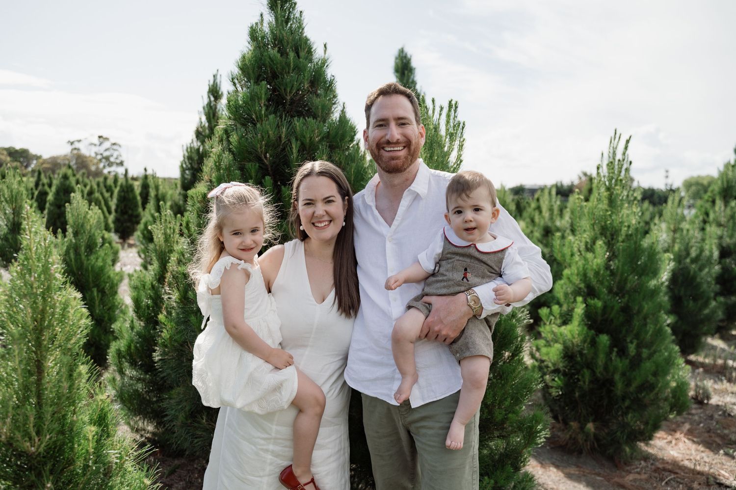 Family of four smiling at a Christmas tree farm, parents holding young children among lush green pine trees.