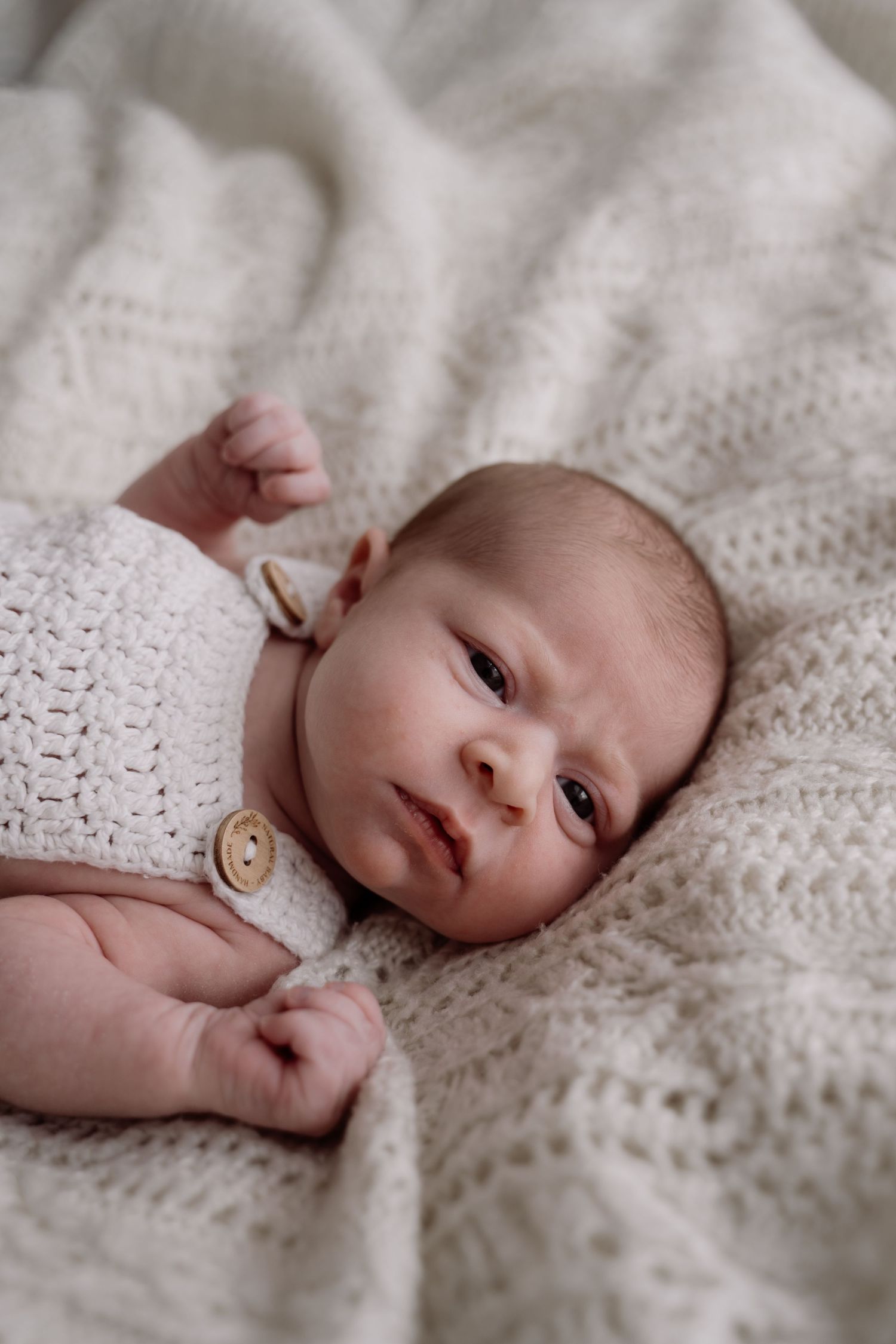 Newborn baby in cream crochet outfit lying on white knit blanket, eyes open, tiny fists raised.