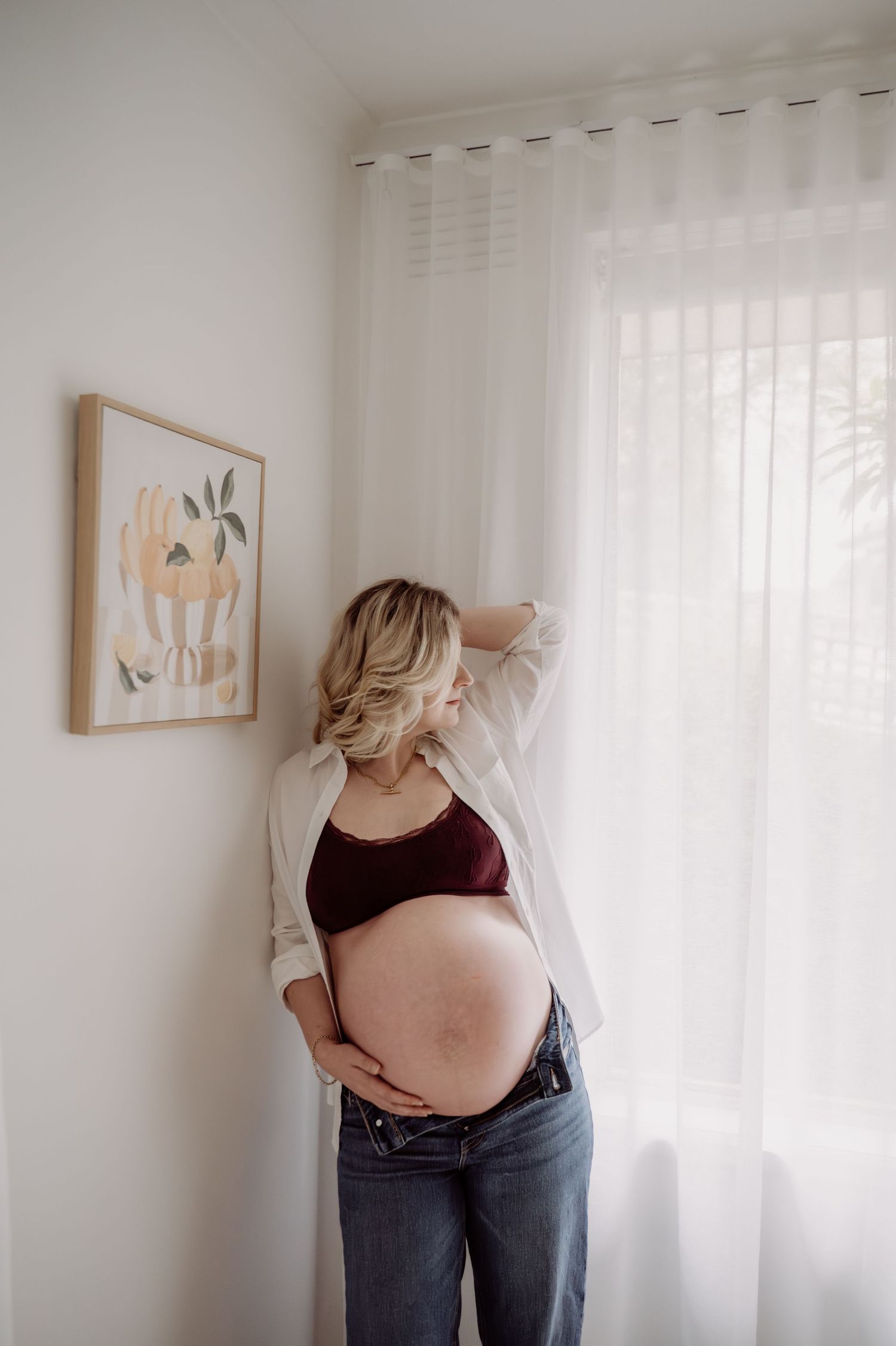Pregnant woman in burgundy crop top and jeans cradling her baby bump near a sunlit window with sheer curtains.