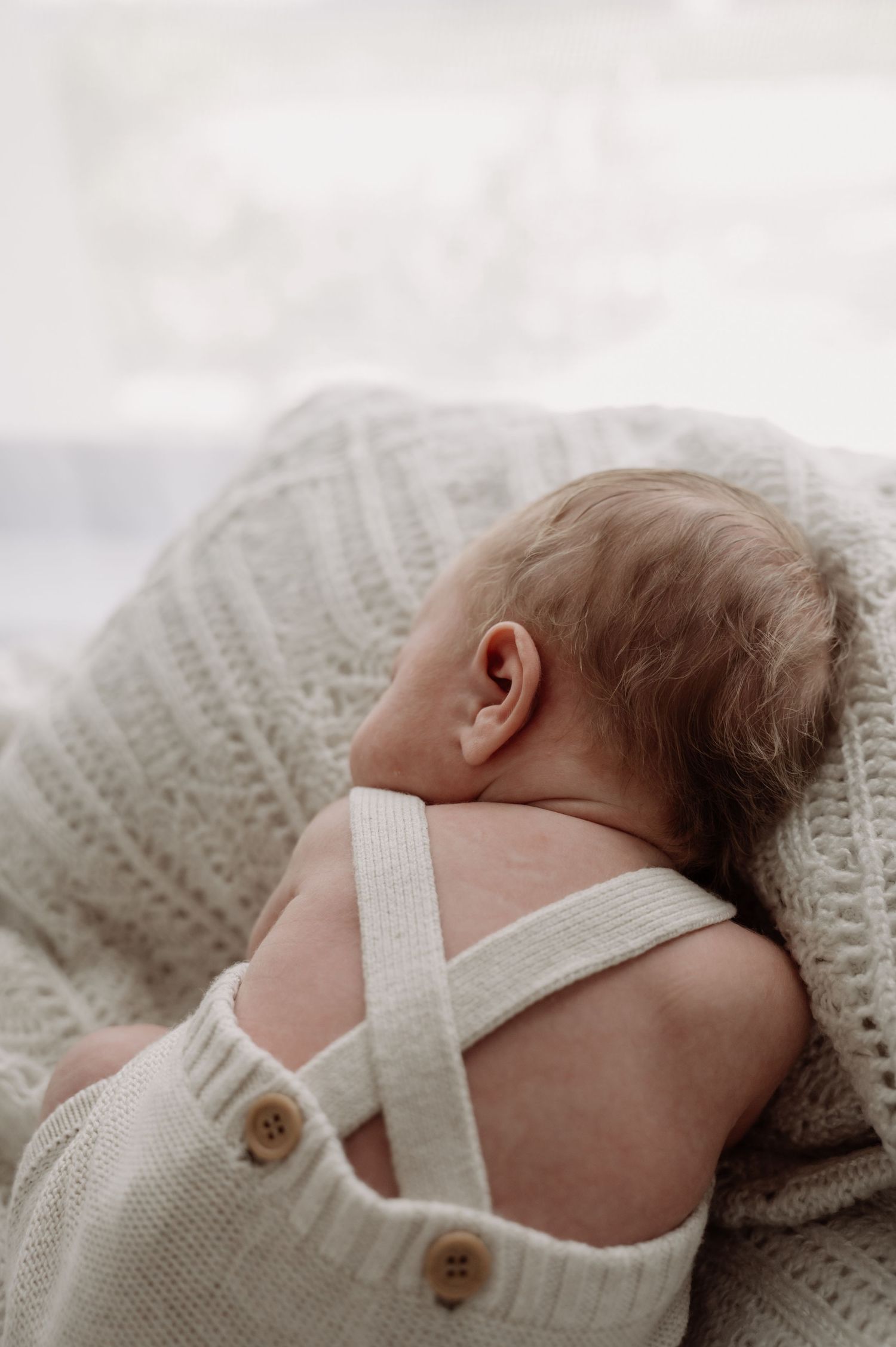 A sleeping newborn baby wearing a cream knit romper with button straps, resting on a soft white knitted blanket.