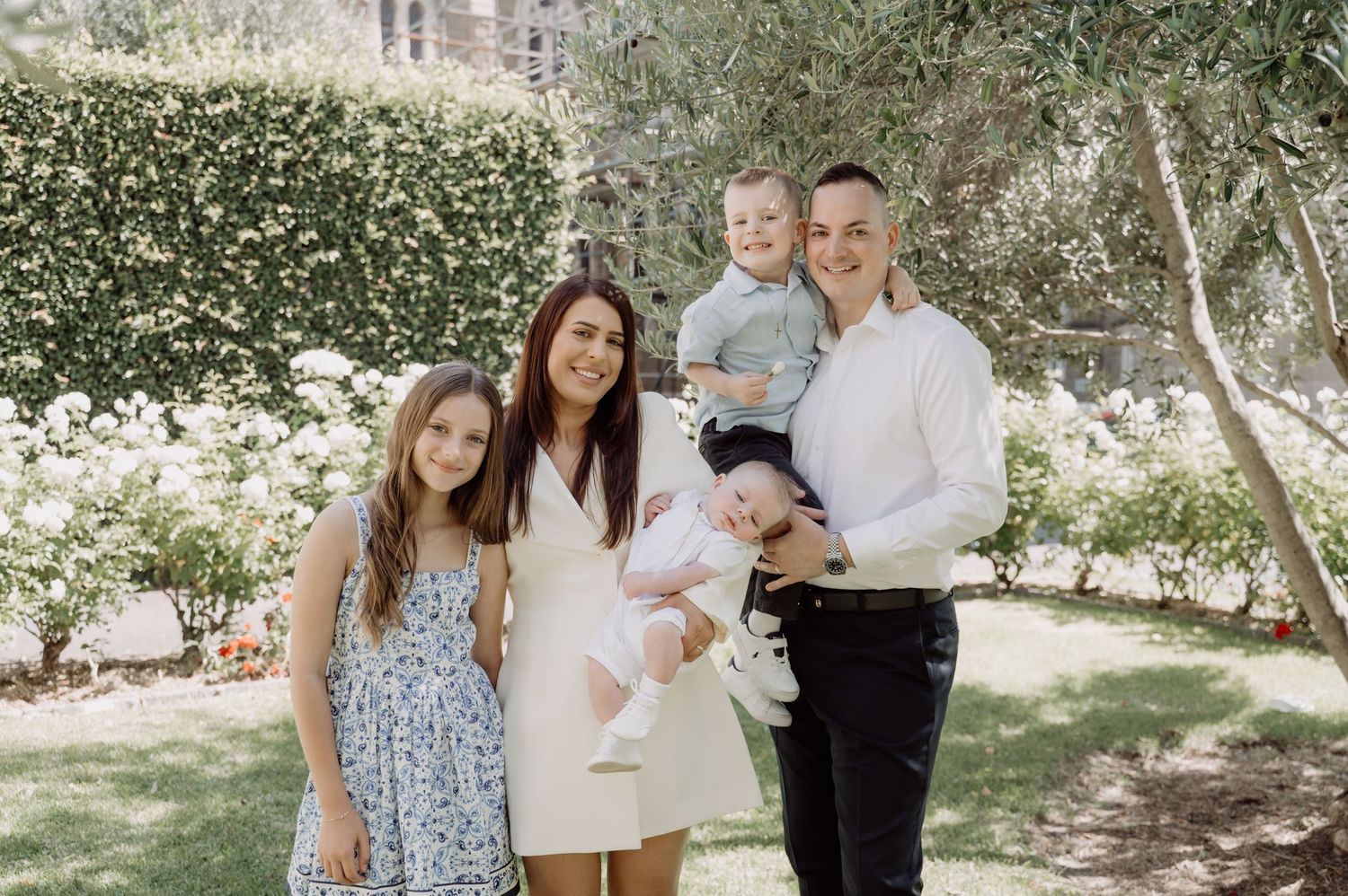 Family of five posing outdoors in a lush garden, parents with three young children smiling for a portrait photo.