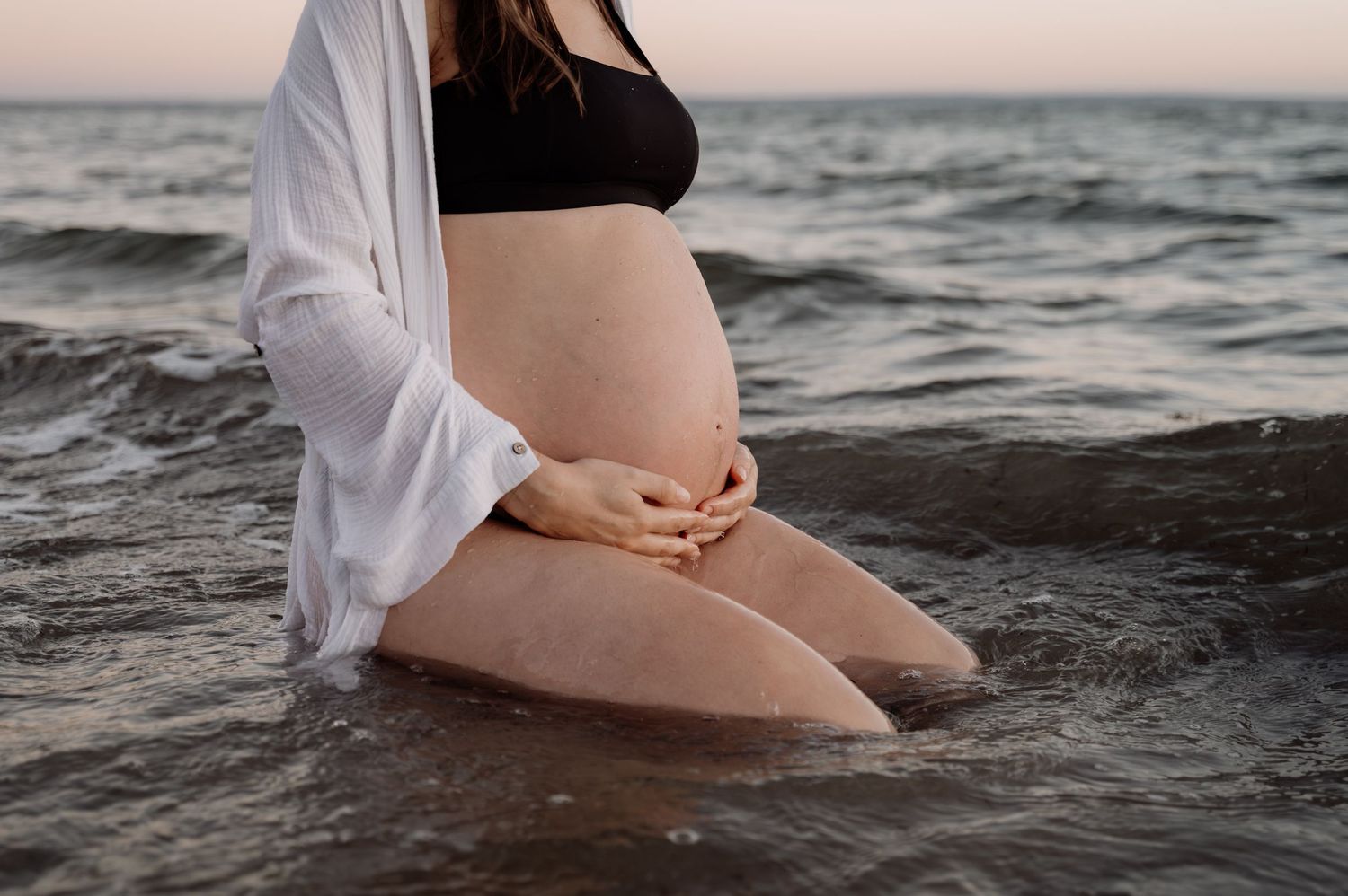 Pregnant woman standing in ocean waves, cradling her baby bump, wearing a black bikini top and open white shirt.