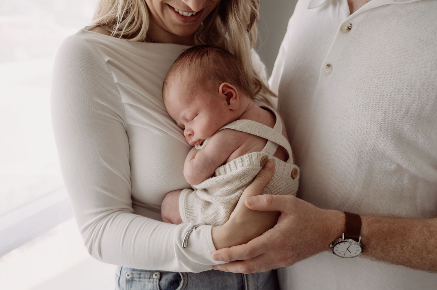 Parents tenderly cradling their sleeping newborn baby, dressed in neutral tones, in a bright and airy setting.