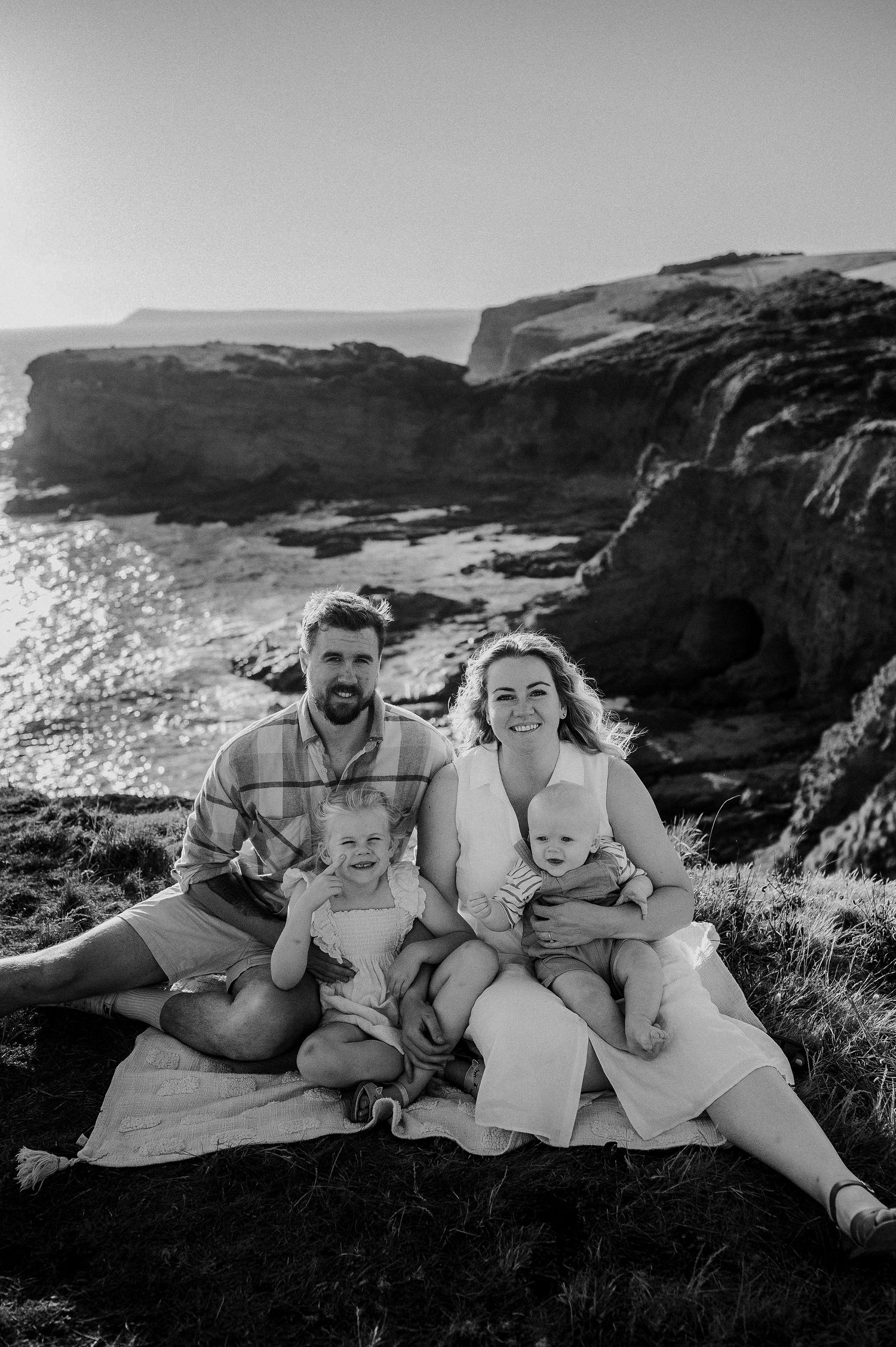 Black and white family portrait on coastal cliffs at sunset with two young children.