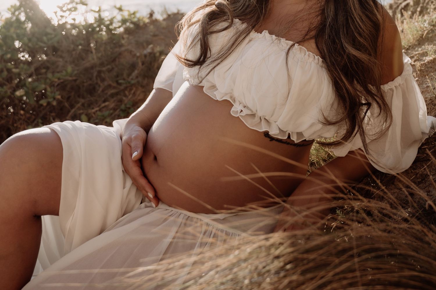 Pregnant woman in white off-shoulder dress sitting in golden field, cradling bare baby bump in warm sunlight.