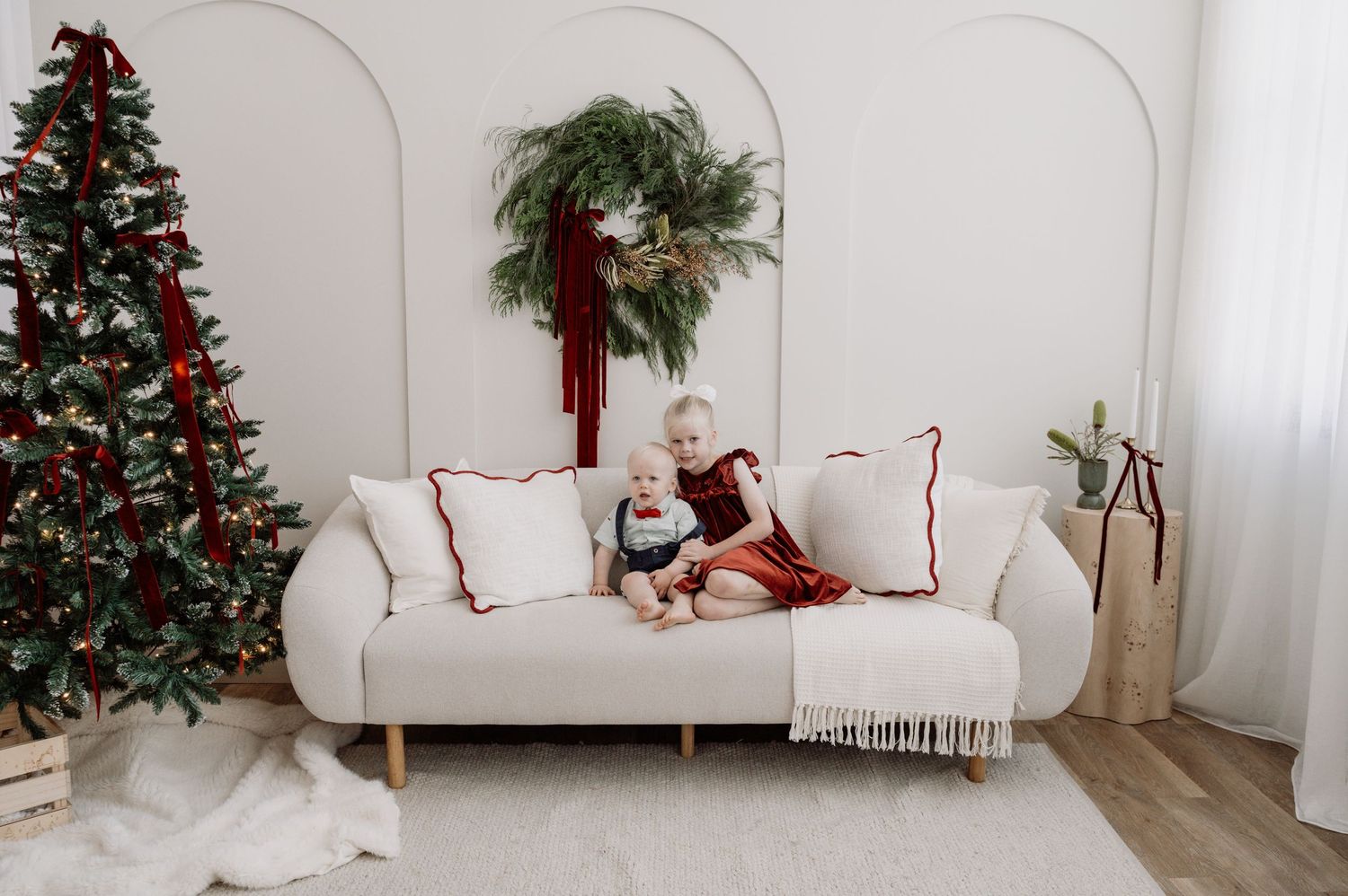 Two young children sit together on a white sofa beside a decorated Christmas tree in a festive holiday living room.