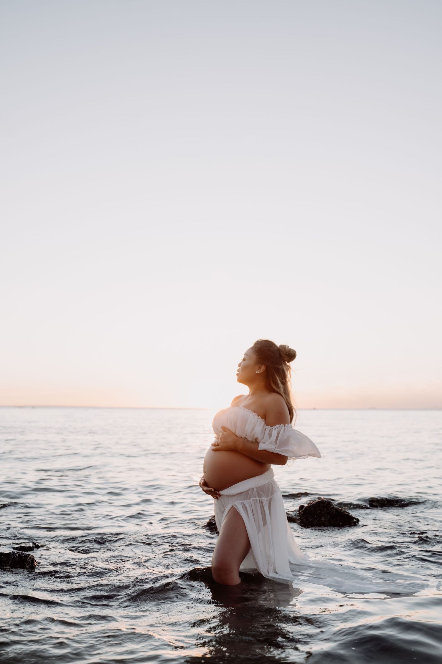 Pregnant woman in white off-shoulder dress standing in ocean at sunset.