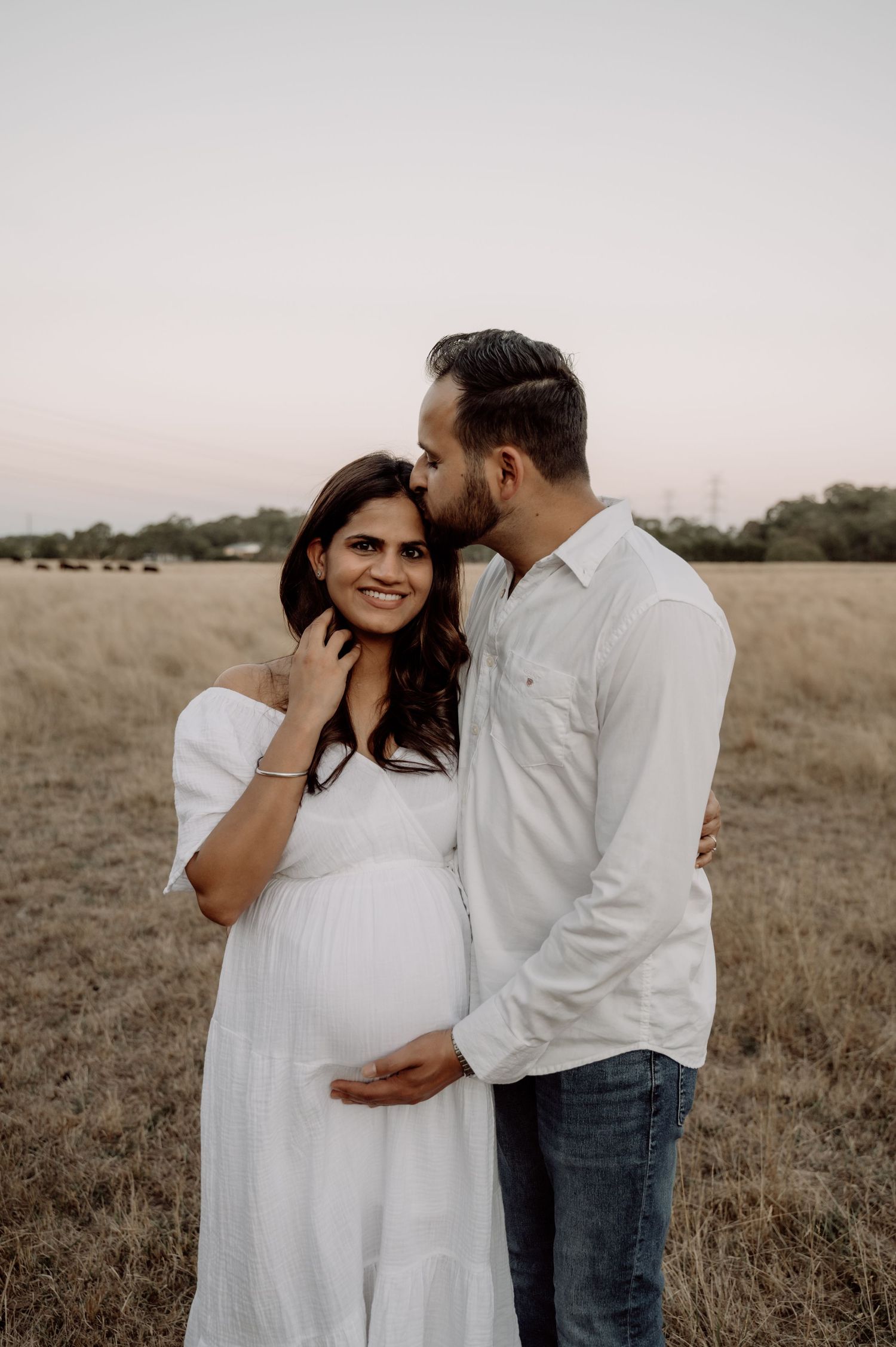 Couple sharing a tender maternity moment in a golden field at sunset, man kissing pregnant woman.