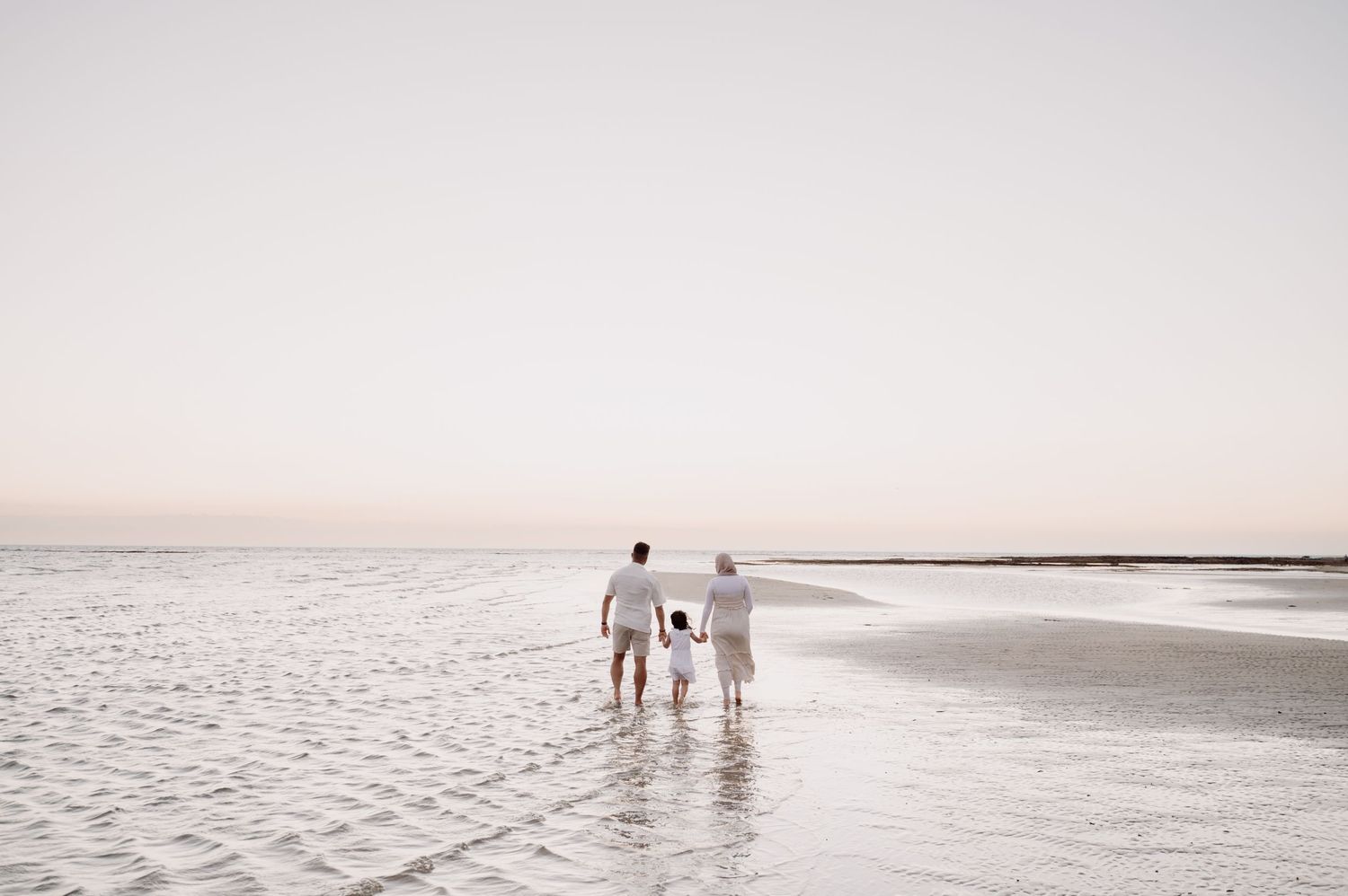 Family of three walking hand-in-hand along a serene, shallow beach at sunset, all dressed in white.