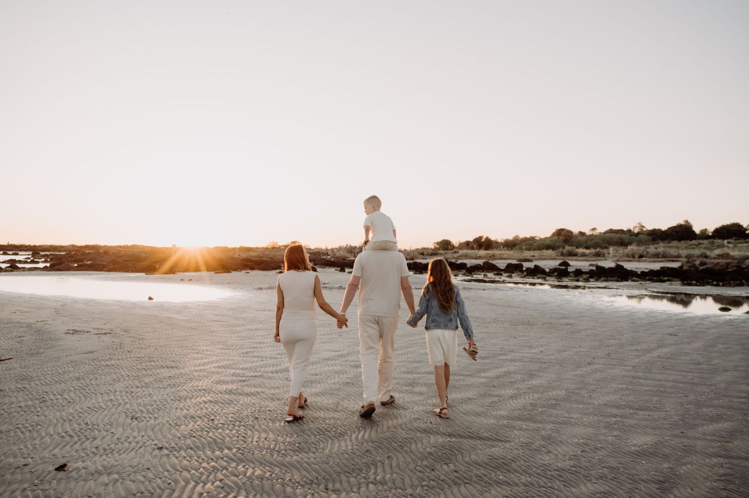 Family walking on beach at sunset, parents holding hands with children, golden light reflecting on sand.