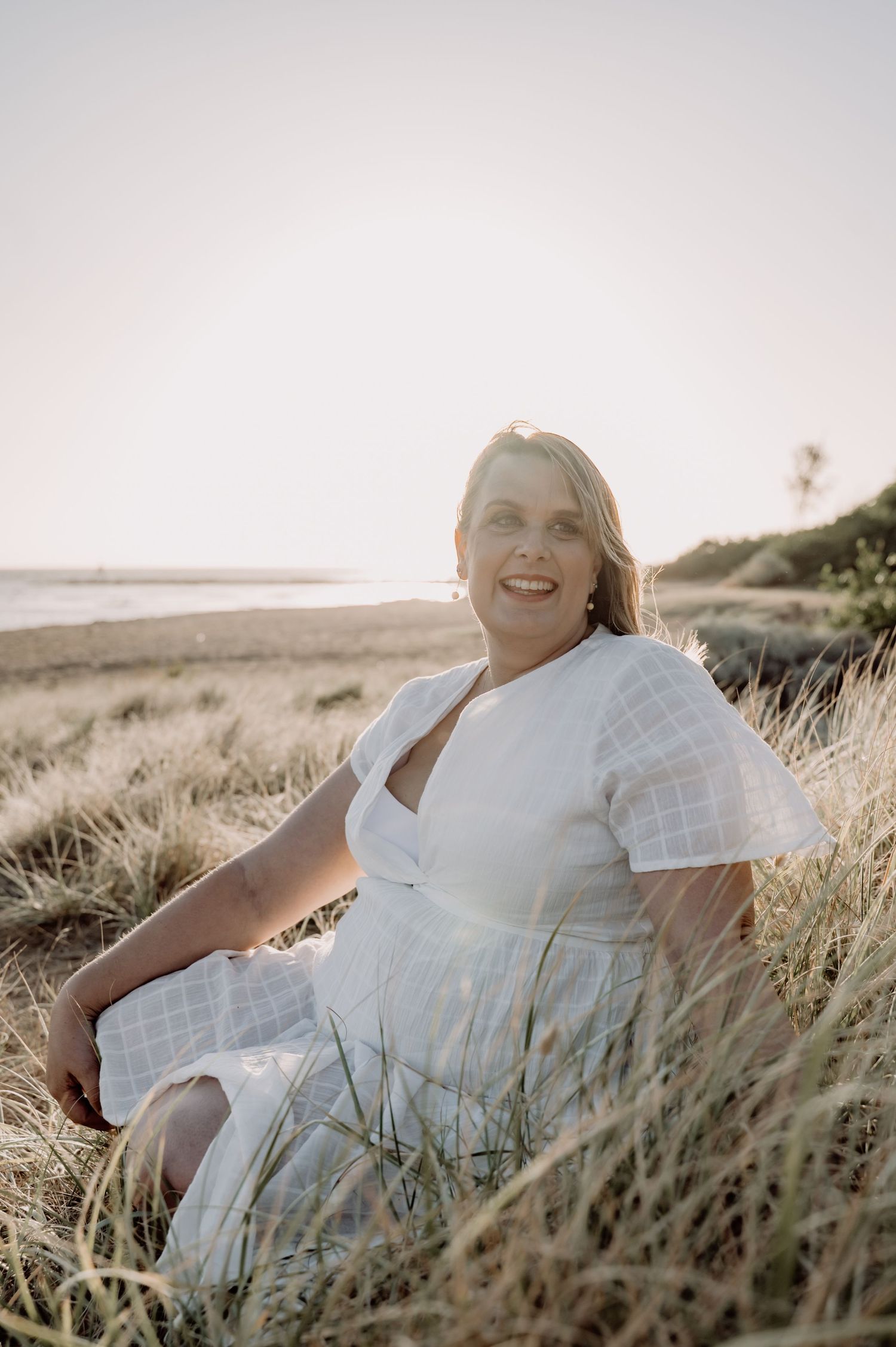 Woman in white dress sitting in coastal dunes at golden hour, smiling warmly at camera.