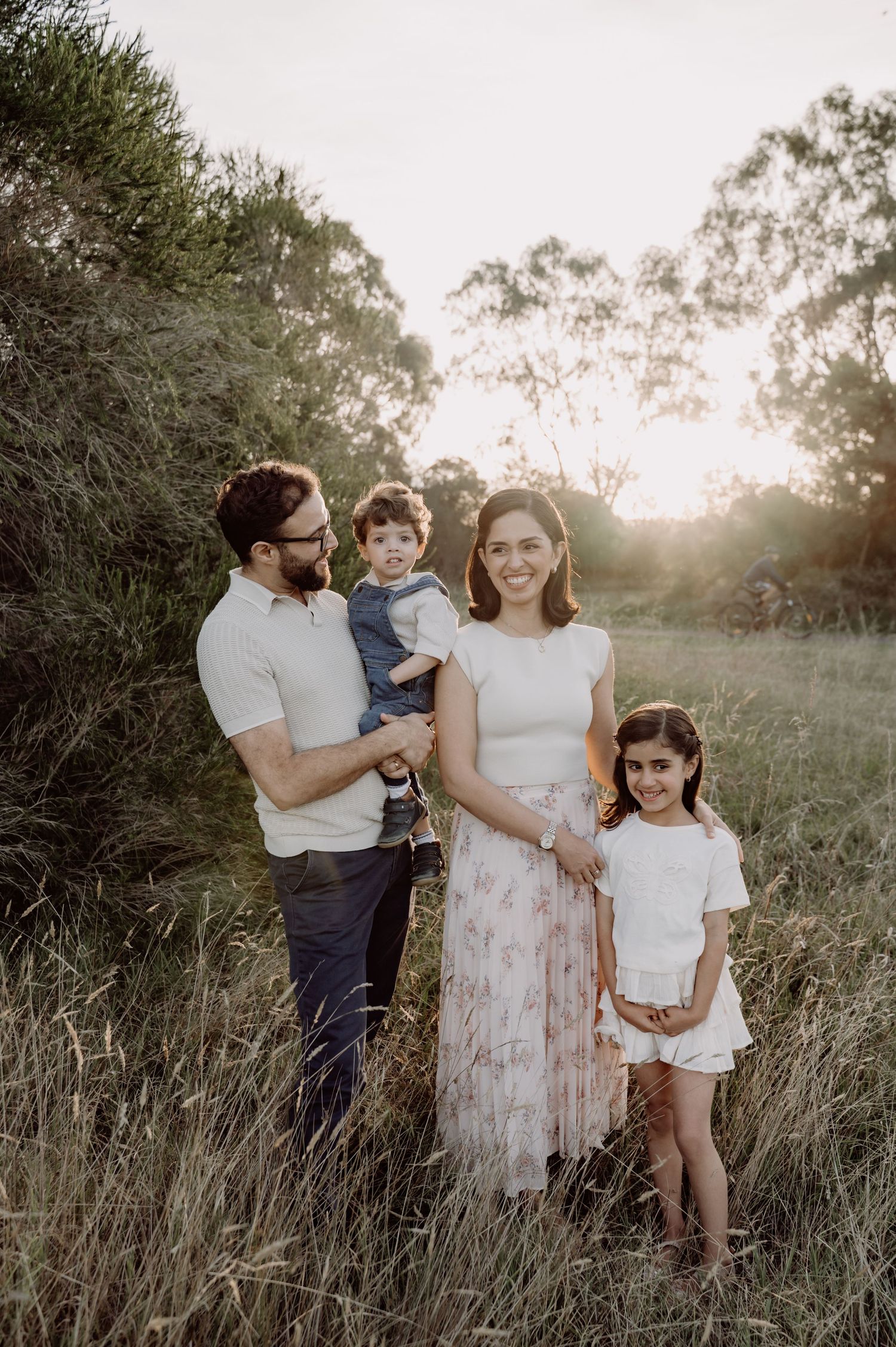 Family of four posing in golden hour sunlit field, parents with young boy and girl surrounded by tall grass and trees.