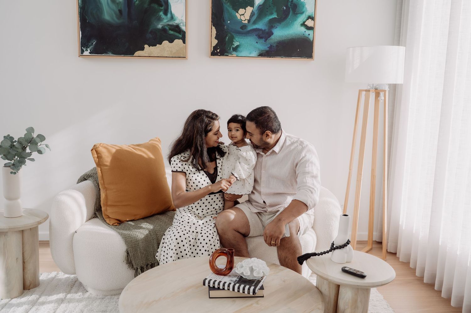 A happy family of three sits together on a white sofa in a bright modern living room with abstract wall art.