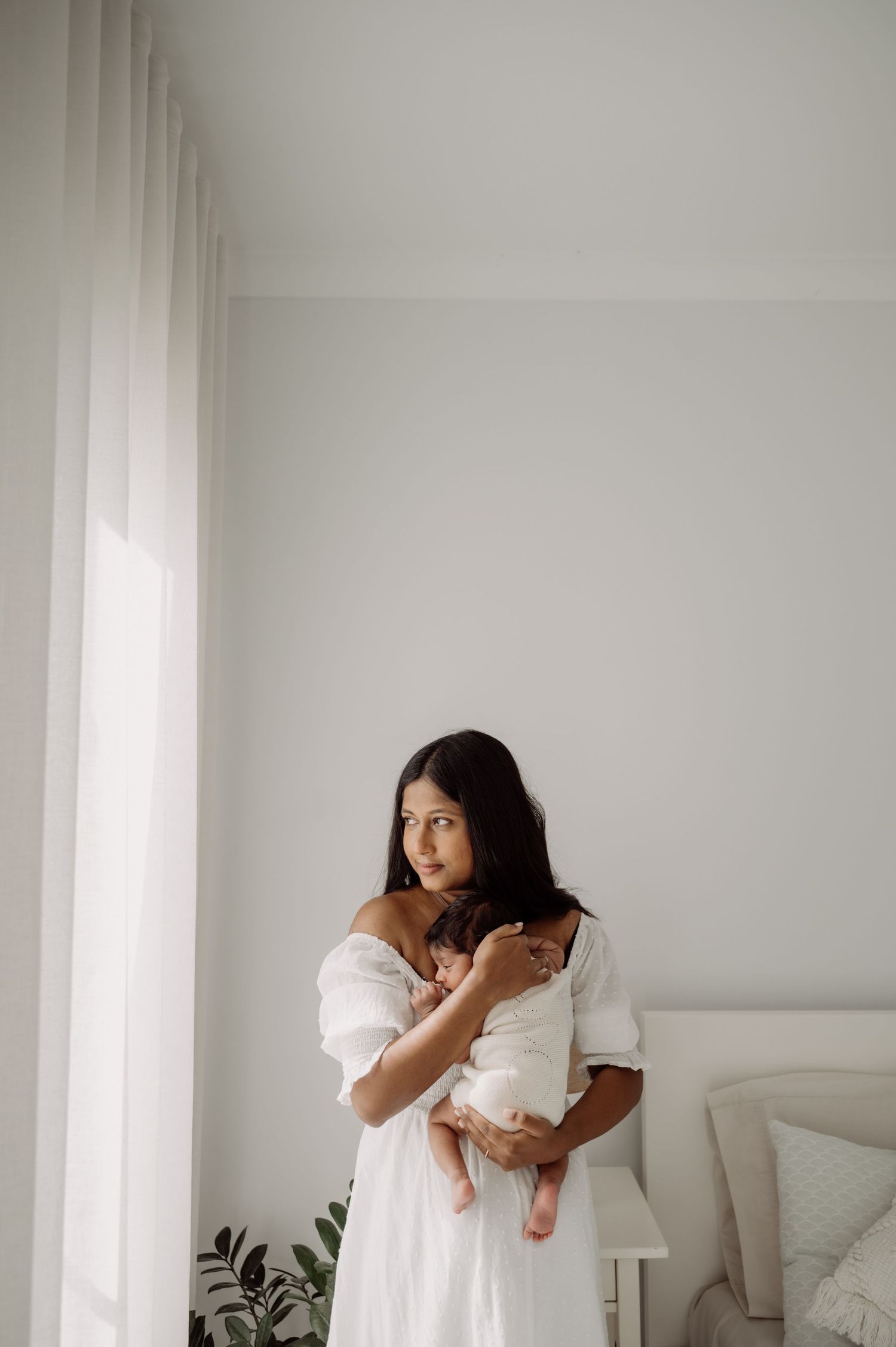 A mother in a white off-shoulder dress tenderly holds her newborn baby near a bright window in a serene white bedroom.