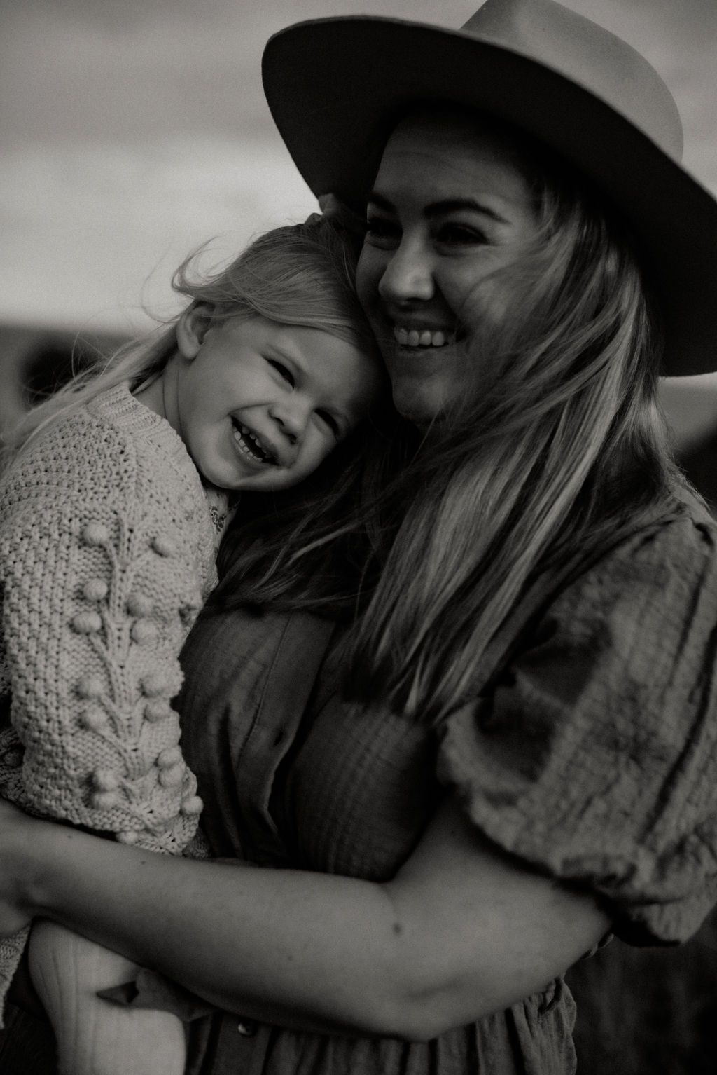 Black and white photo of laughing mother in hat hugging smiling young daughter.