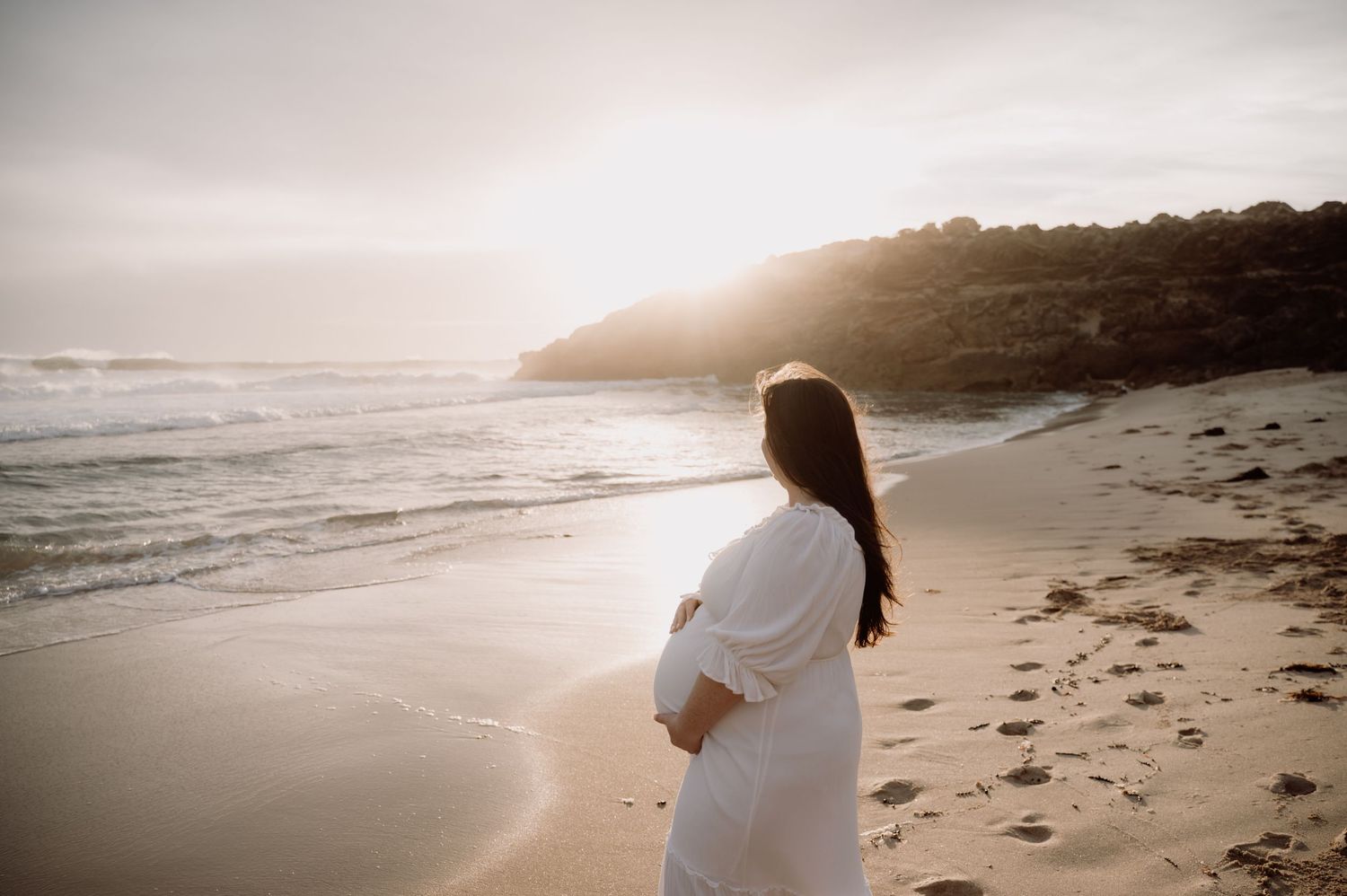 Pregnant woman in white dress standing on sandy beach at sunset, cradling her baby bump while gazing at ocean waves.