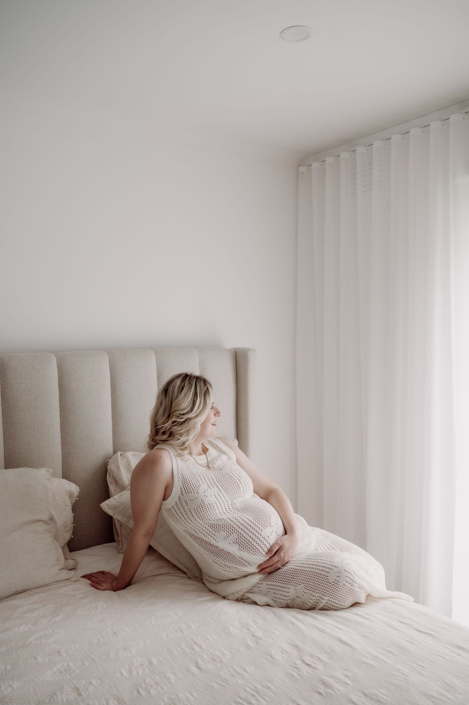 Pregnant woman in white dress sitting on bed, cradling baby bump, soft natural light, minimalist bedroom.