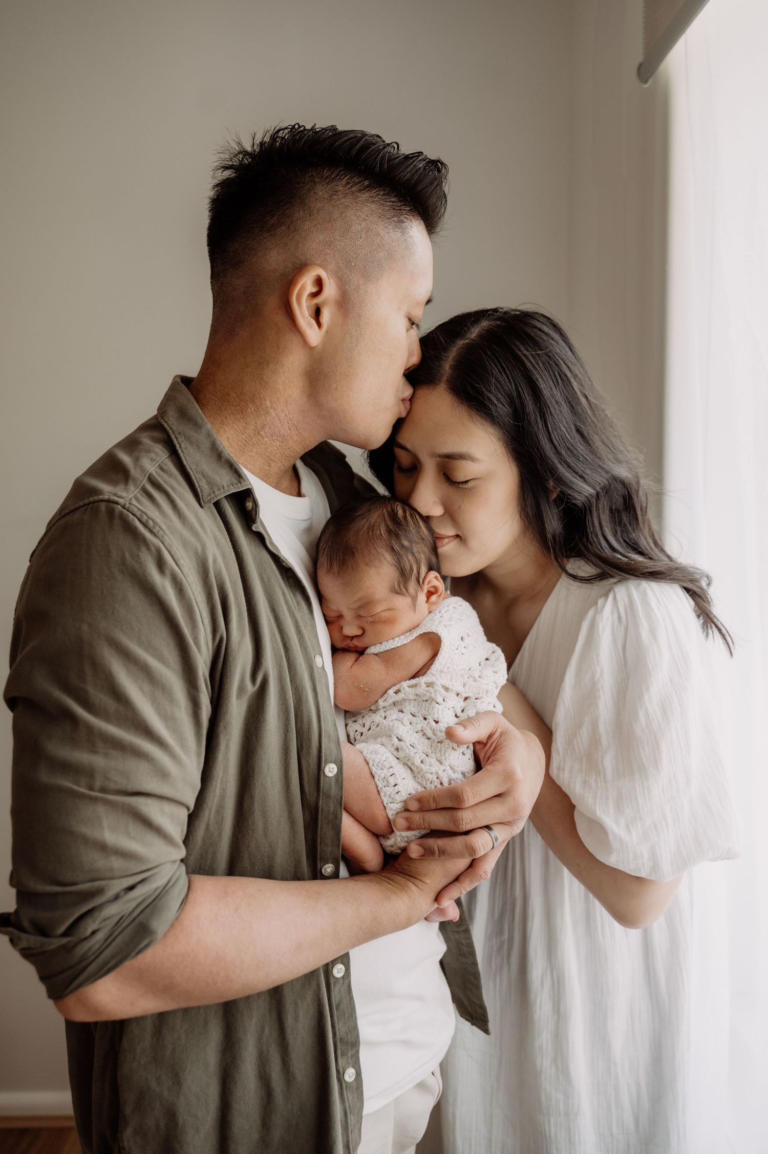 Asian couple tenderly kissing their sleeping newborn baby by a bright window.