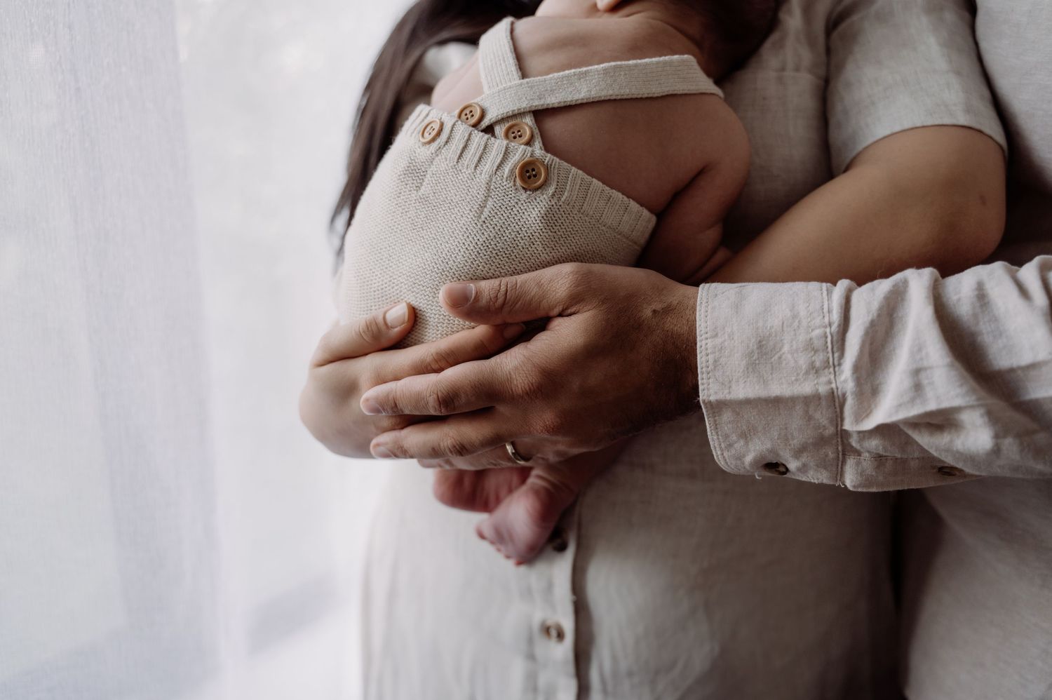 A mother tenderly holds her newborn baby dressed in a neutral linen romper near a bright window.