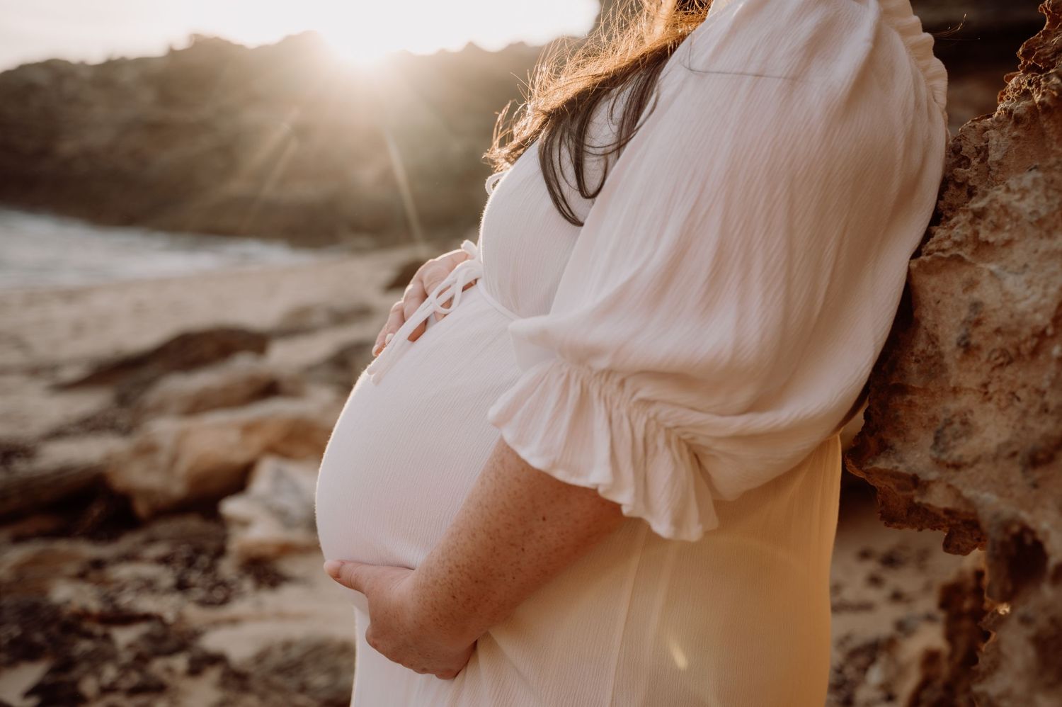 Pregnant woman in white dress cradling baby bump at golden hour on rocky beach.