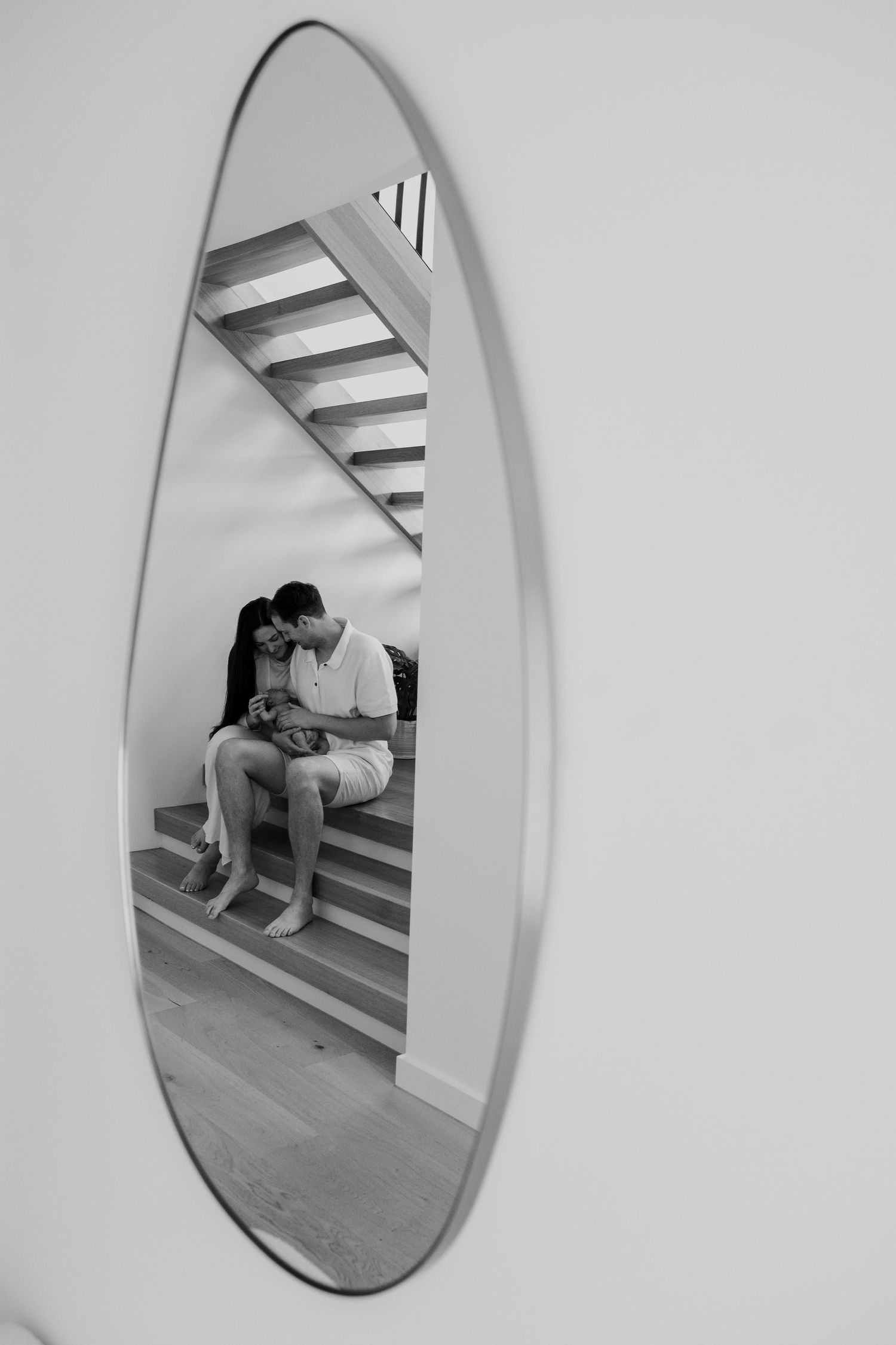 Black and white reflection of couple with newborn in oval mirror on modern staircase.