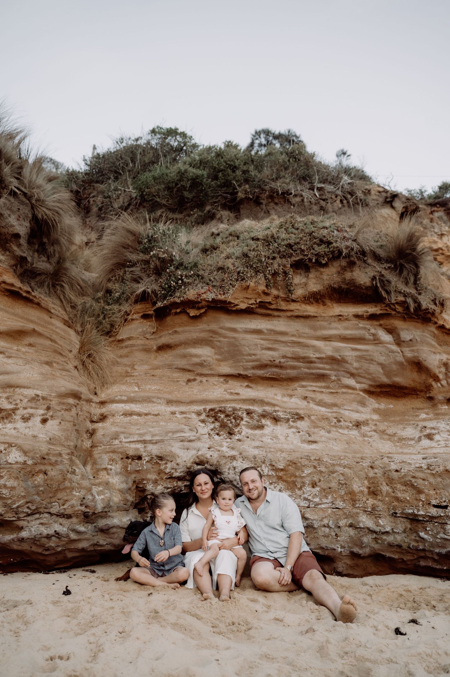 Family of four sitting together against red sandstone cliffs on a beach.