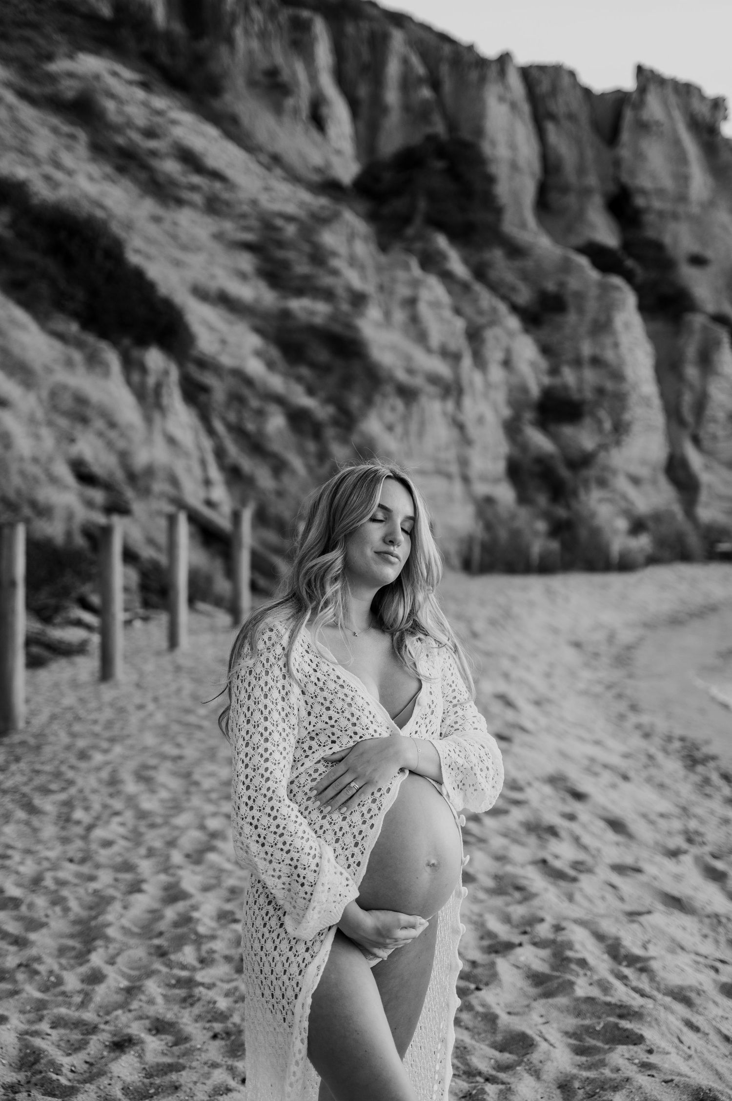 Black and white maternity photo of pregnant woman in crochet wrap posing on beach with cliff backdrop.