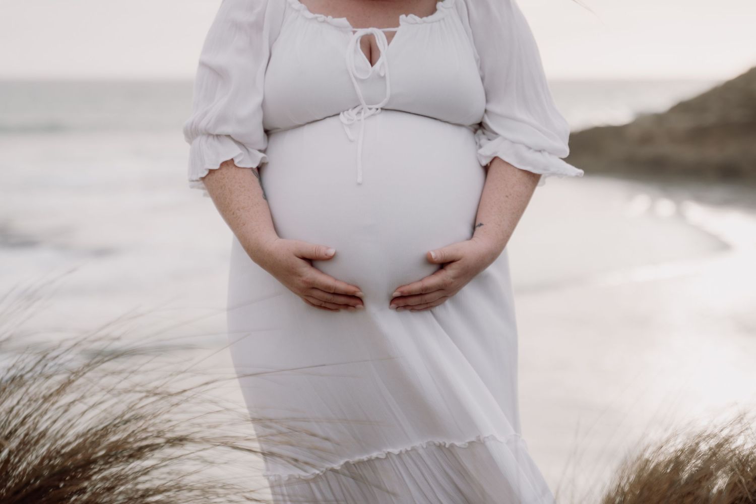 Pregnant woman in white dress cradling baby bump on beach with ocean and sand dunes in background.