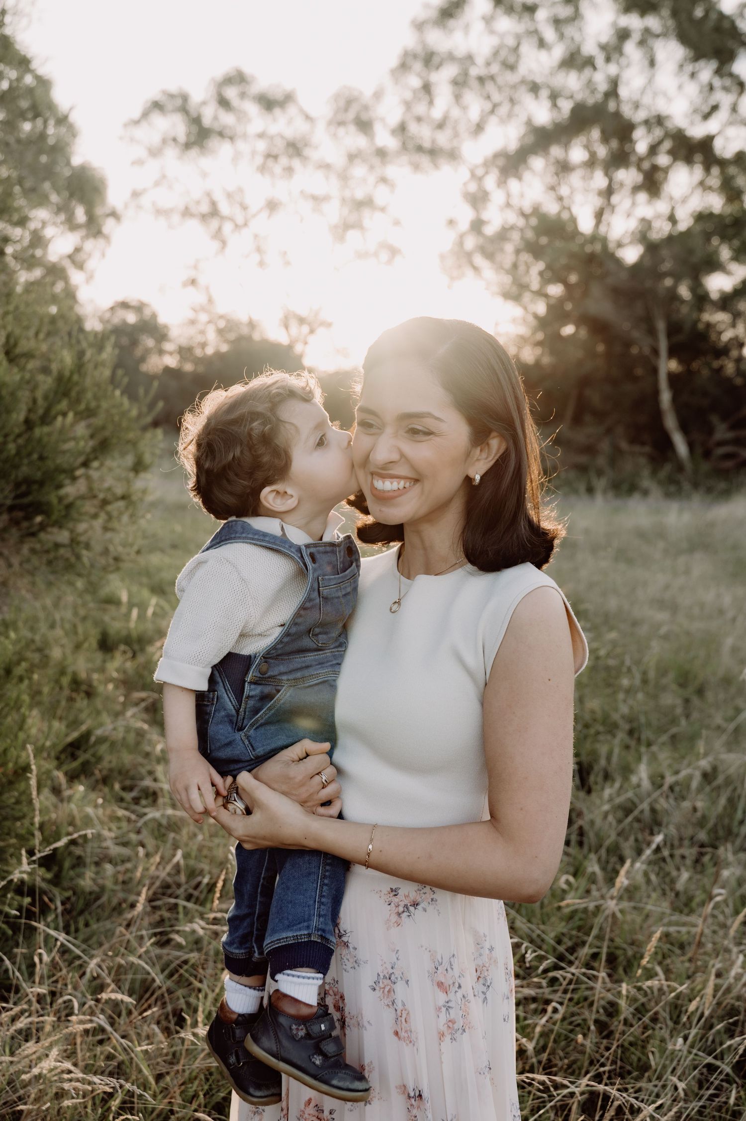 A smiling woman holds a toddler boy who kisses her cheek in a sunlit outdoor field.