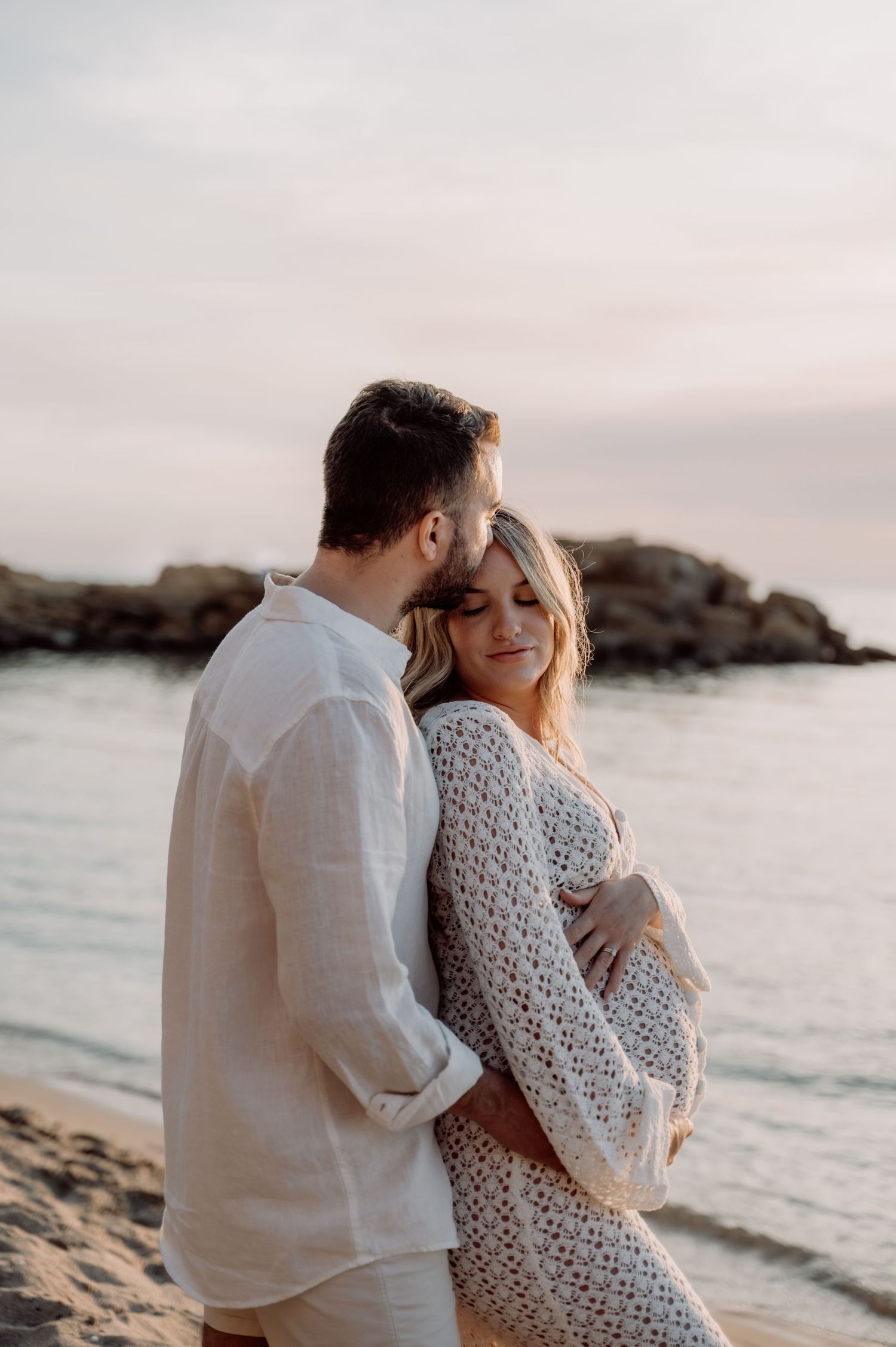 Couple embracing on beach at sunset, pregnant woman in crochet dress held tenderly by partner near rocky shoreline.