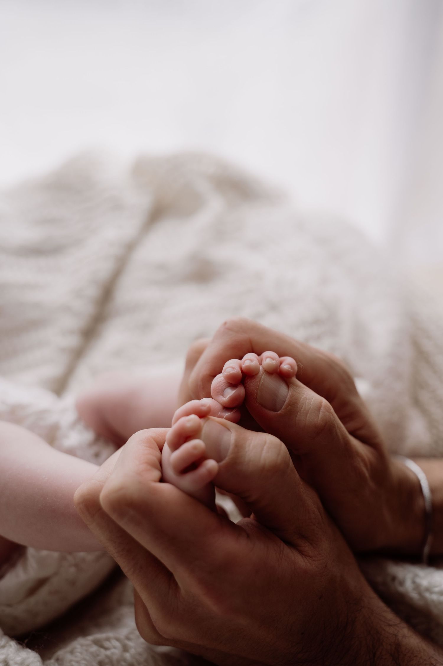 Adult hands gently cradling tiny newborn baby feet on a soft white blanket, close-up maternity photo.