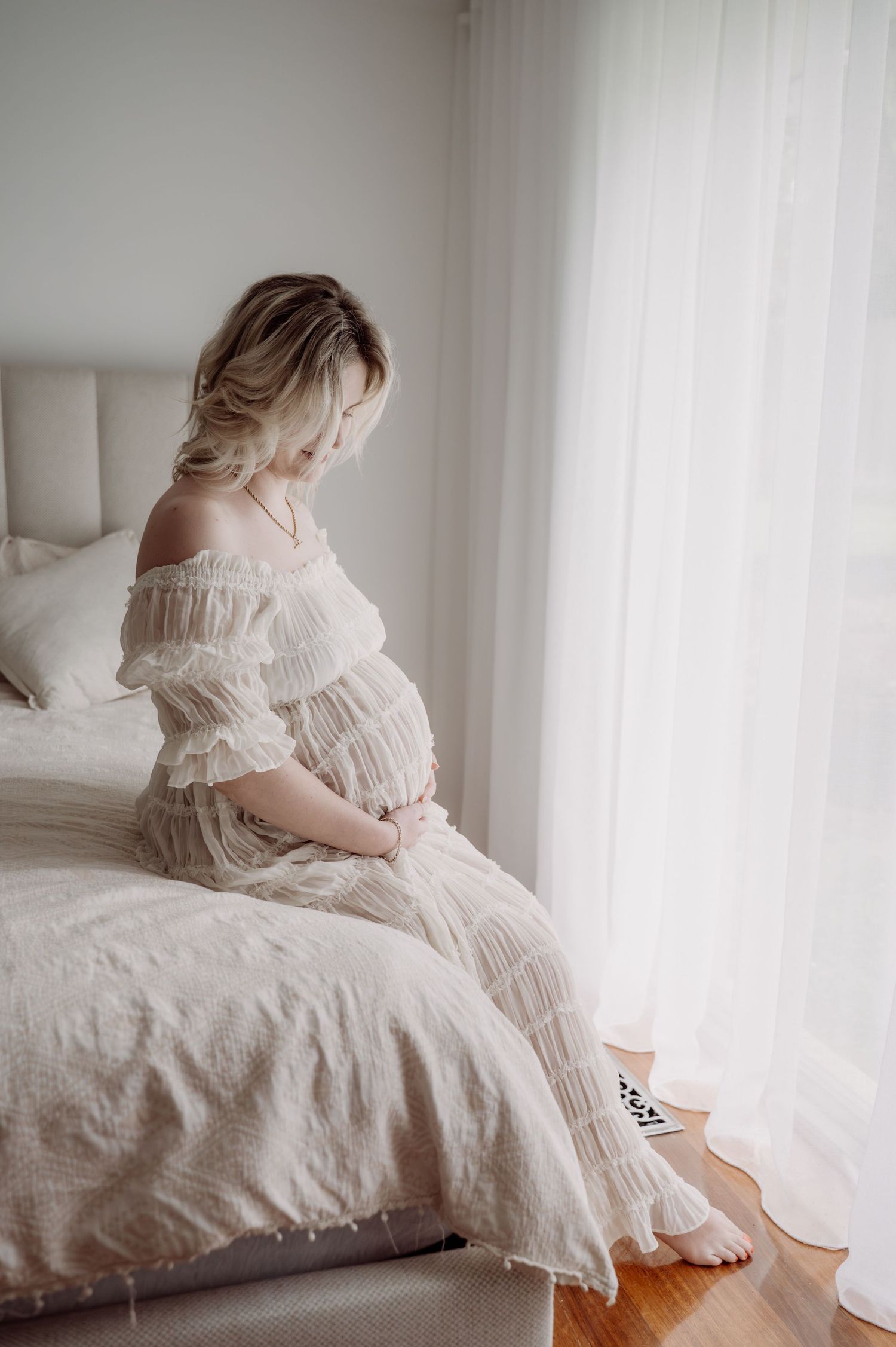 Pregnant woman in white off-shoulder dress sitting on bed near window, cradling baby bump in soft natural light.