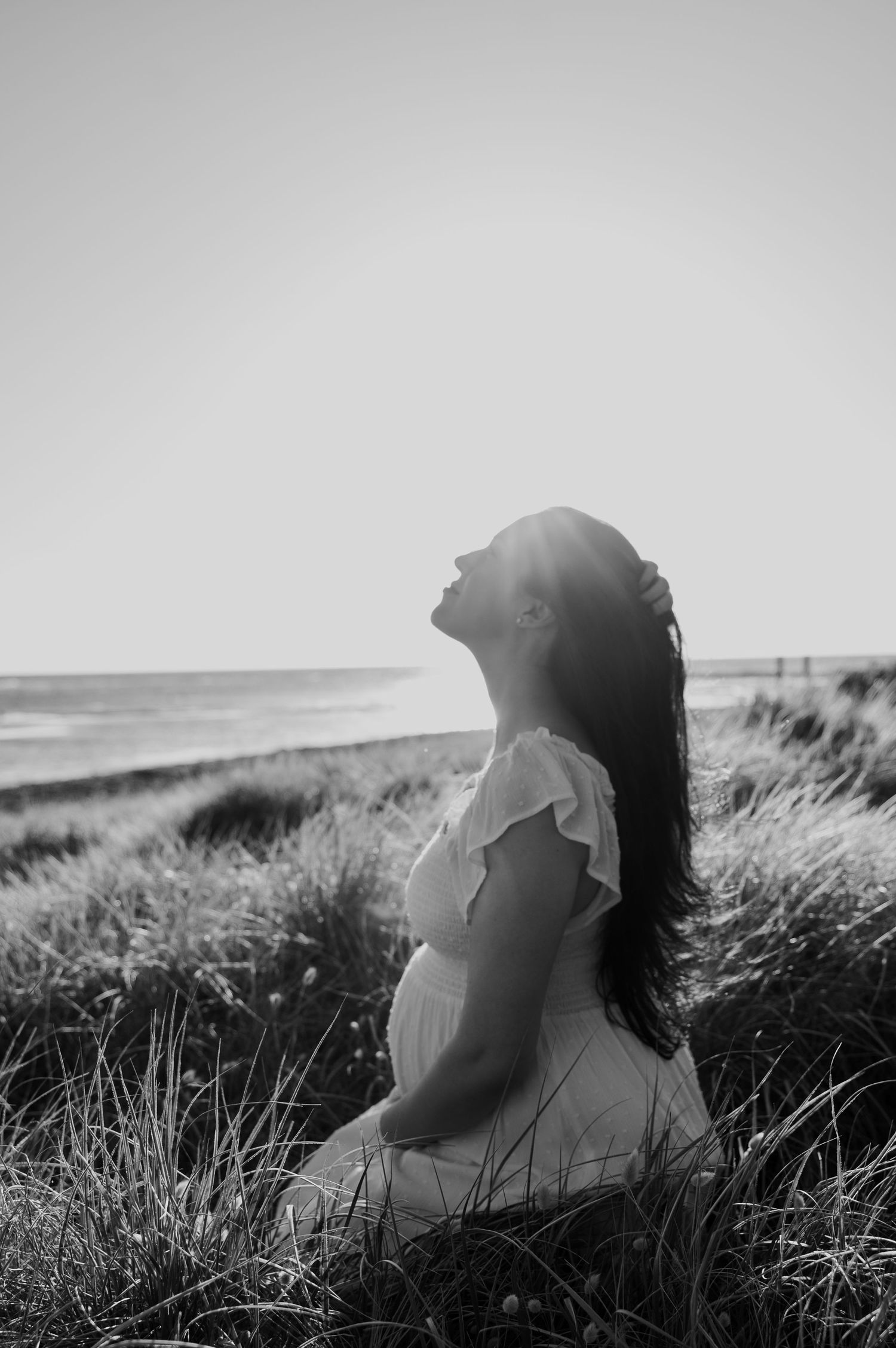 Black and white silhouette of pregnant woman in white dress sitting in beach grass at sunset.